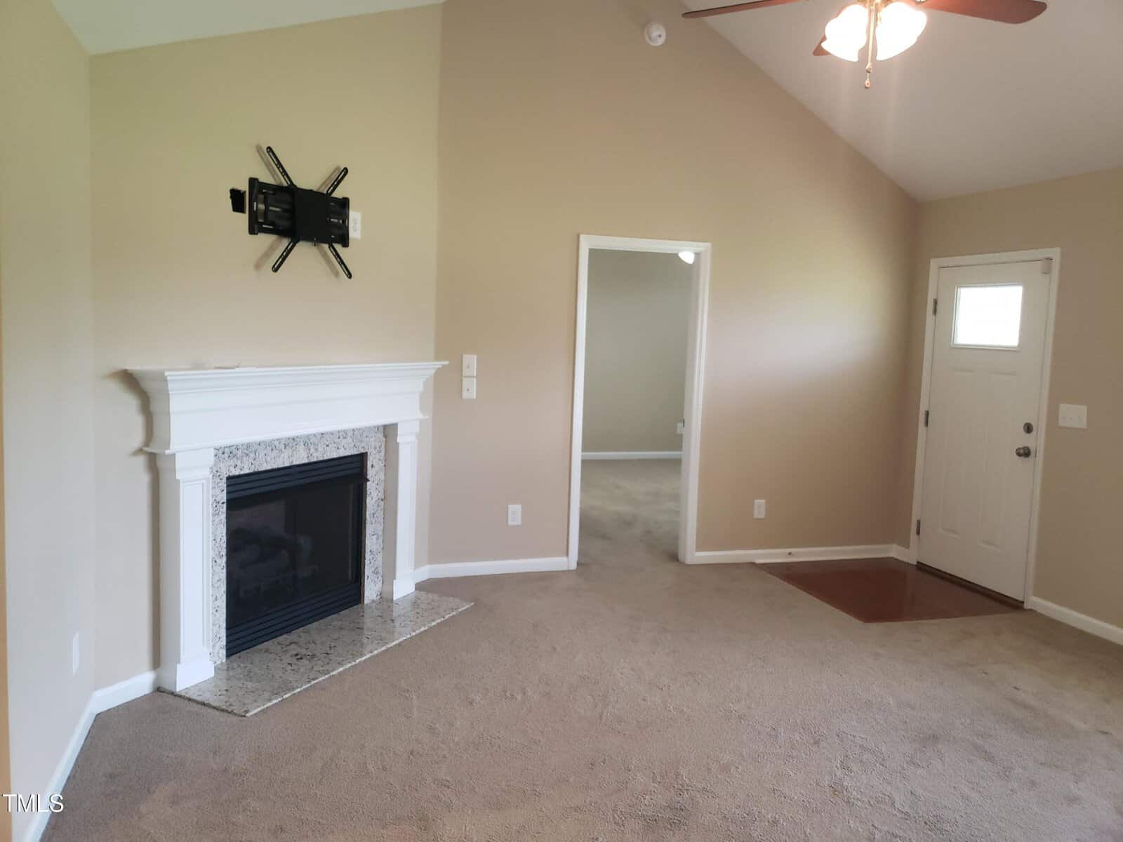 A living room with beige walls features a white fireplace, an empty TV mount above it, and a ceiling fan. There's a doorway leading to another room and a door with a window nearby. The carpet is light brown.