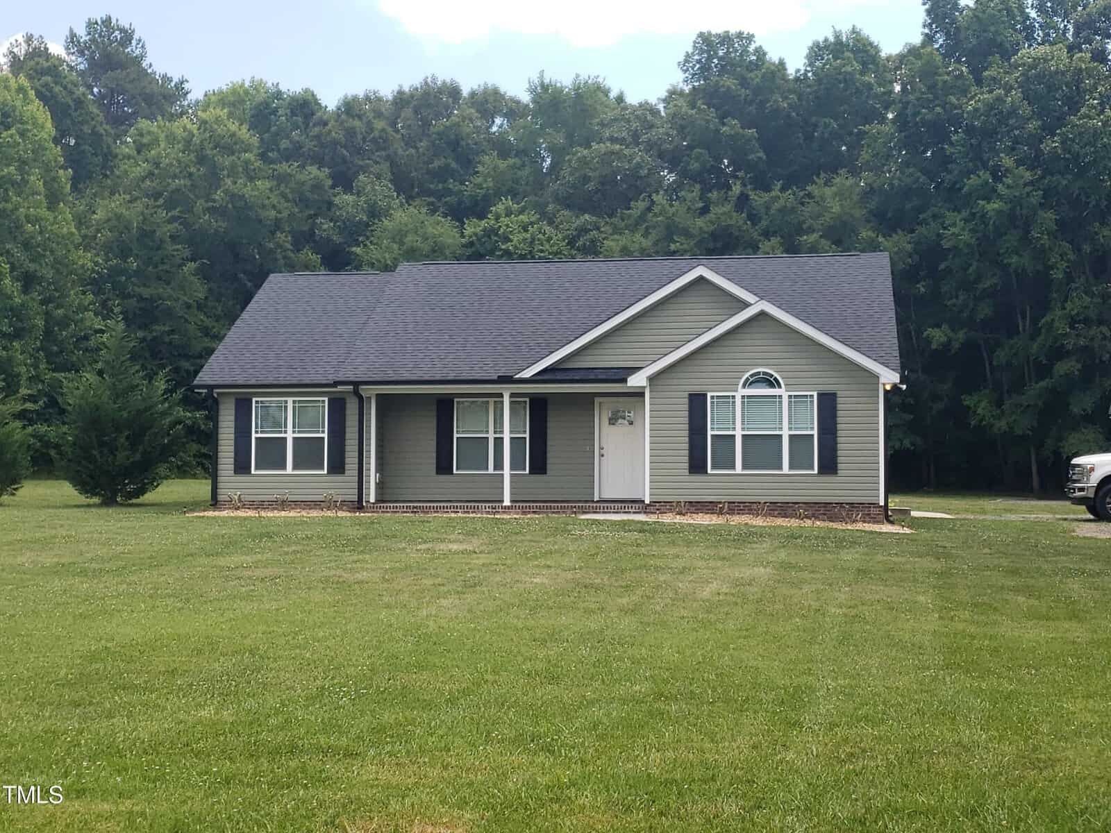 A single-story house with gray siding and a dark roof sits on a large, grassy lawn at 4530 Shock Overton Road. It features a small porch and two front windows. A white vehicle is partially visible on the right, while tall trees form a dense backdrop in Oxford.