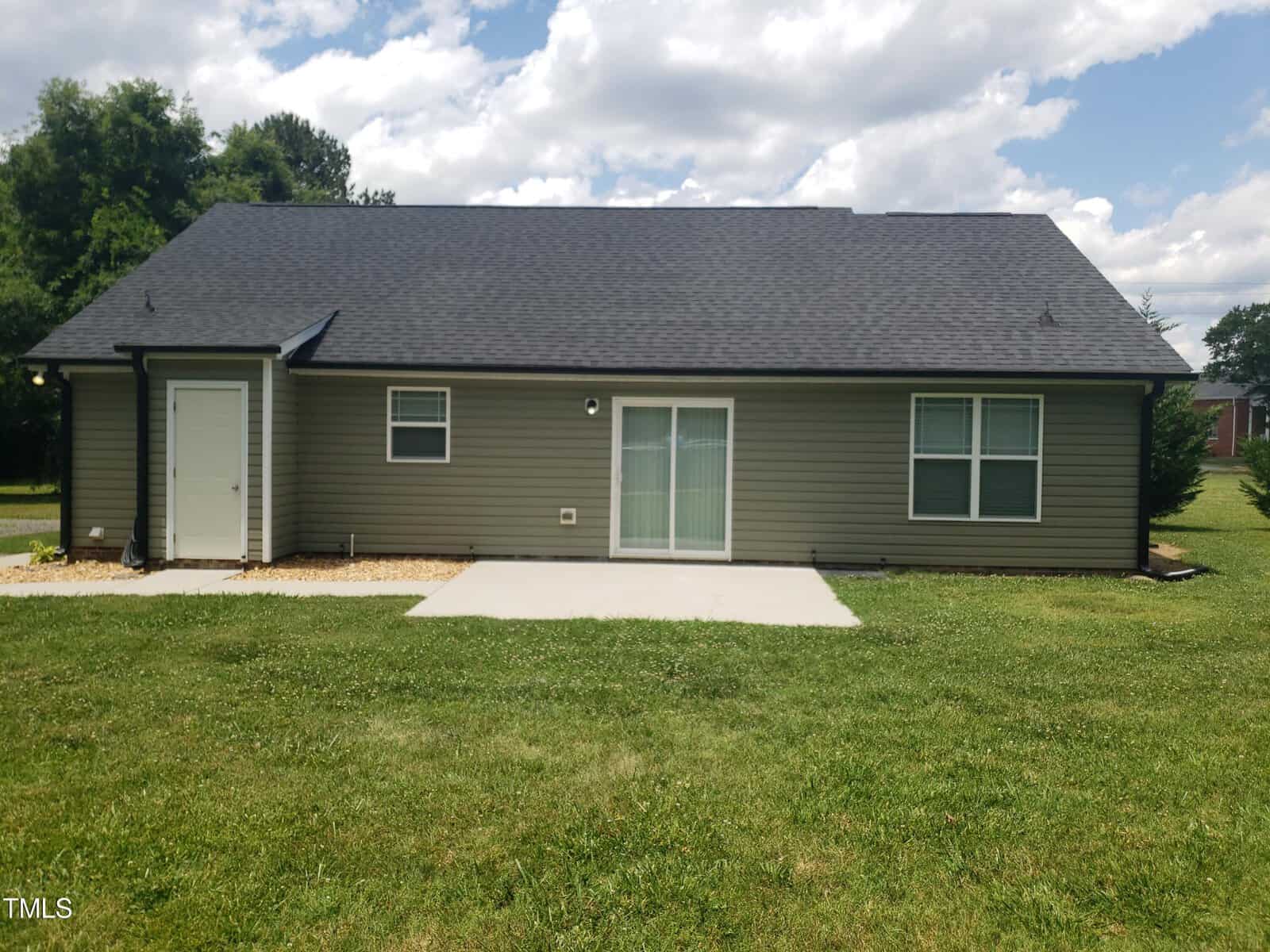 A small, single-story house with a dark roof and light-colored siding. The house has a back door with a small patio and a grassy backyard. The sky is partly cloudy.