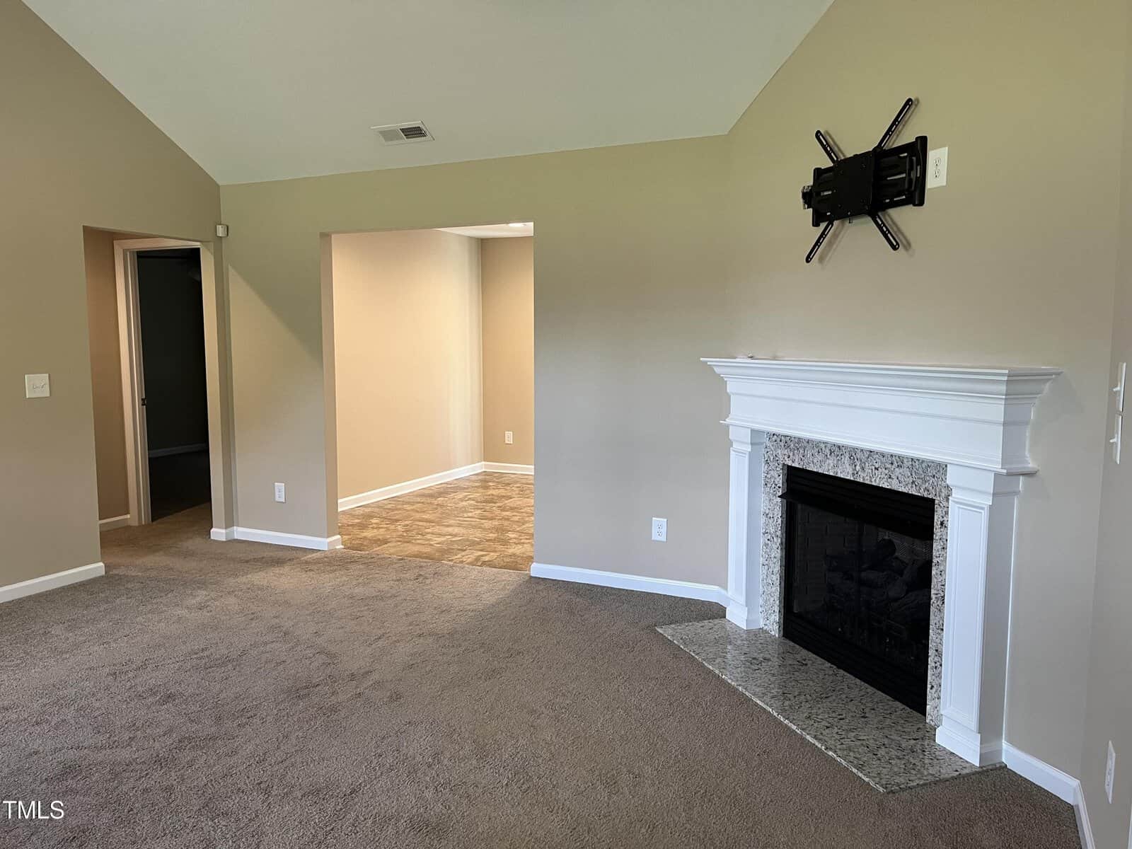 A living room with beige walls and carpet, featuring a white fireplace with a granite hearth. A TV mount is above the fireplace. An open doorway leads to another room with tiled floors. The ceiling is vaulted.