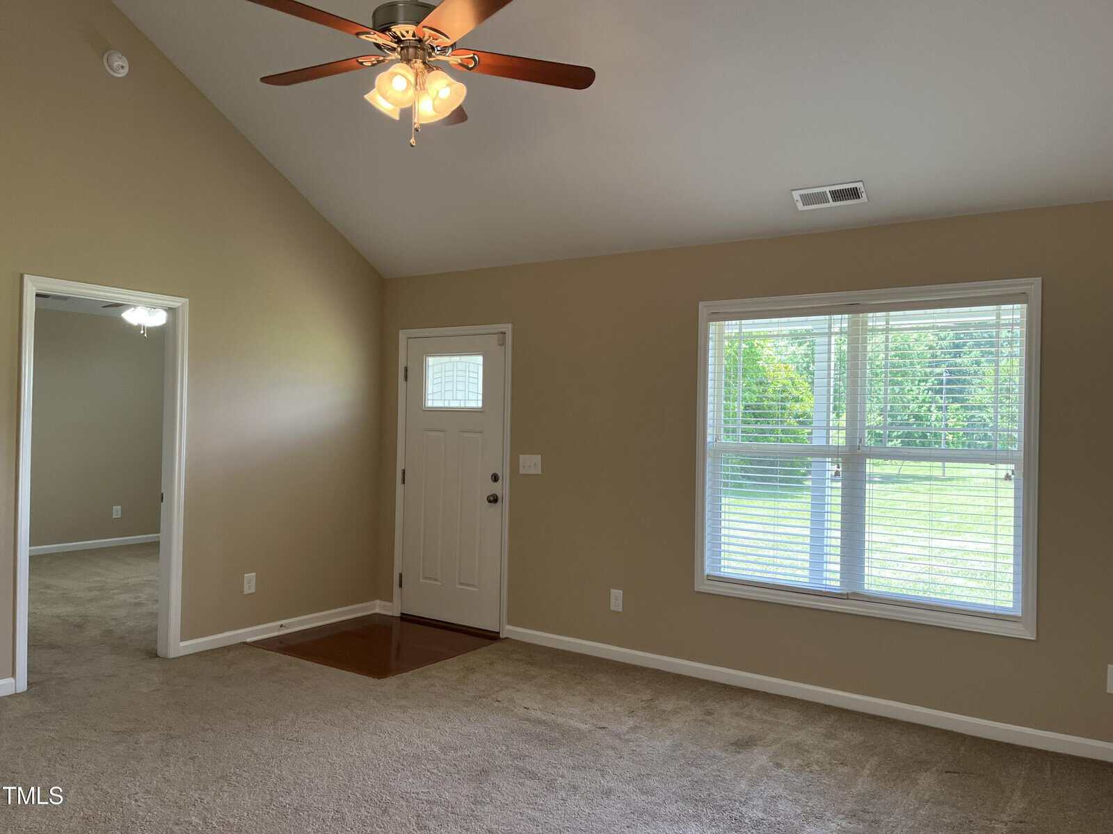 A room with beige walls and carpet, featuring a ceiling fan with lights. There is a white door and a large window on one wall, allowing natural light in. An adjoining room is visible through an open doorway.