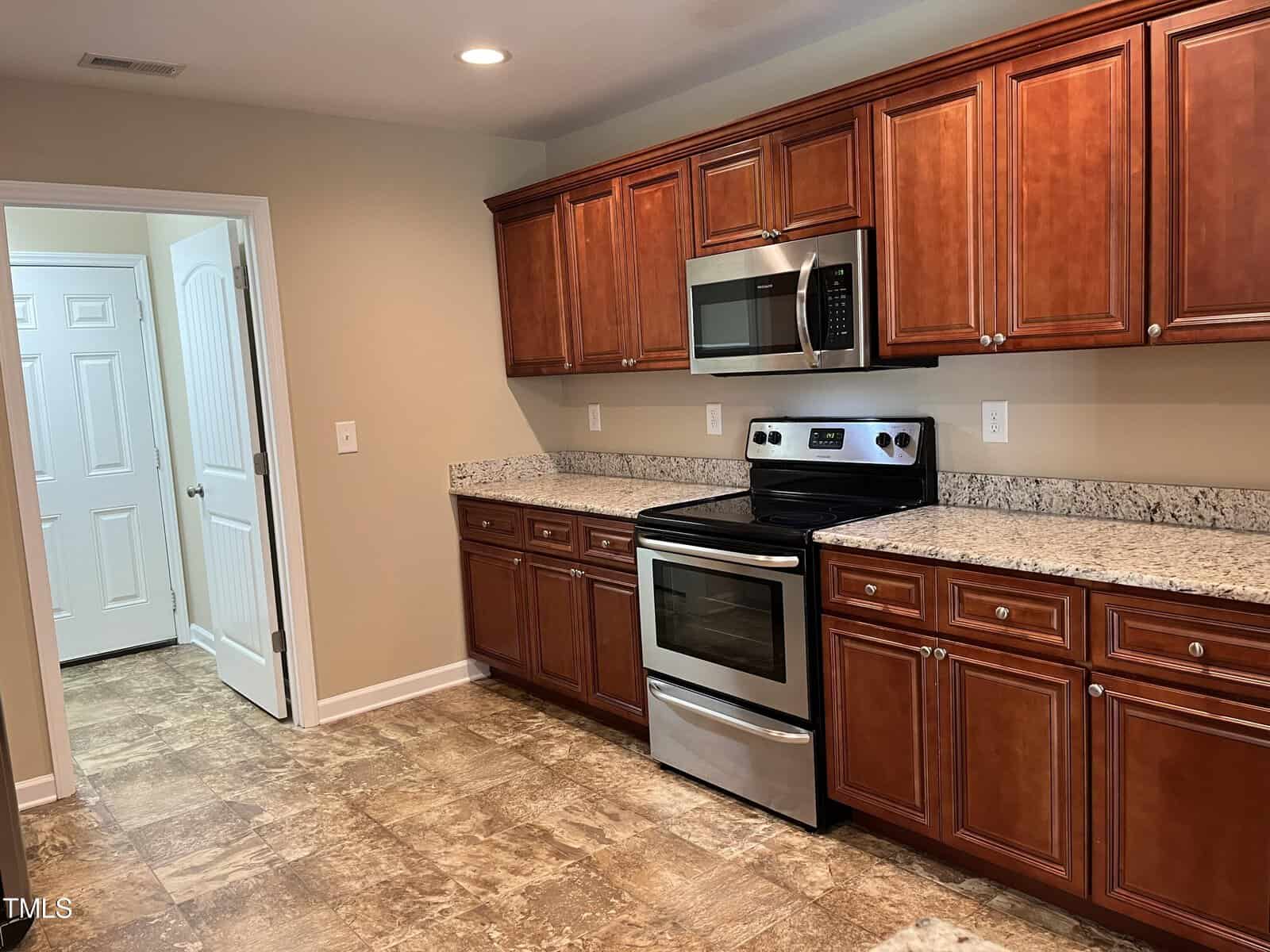 A kitchen with brown wooden cabinets, a granite countertop, and stainless steel appliances, including a microwave and stove. The floor has a tiled pattern, and there's an open door leading to another room.