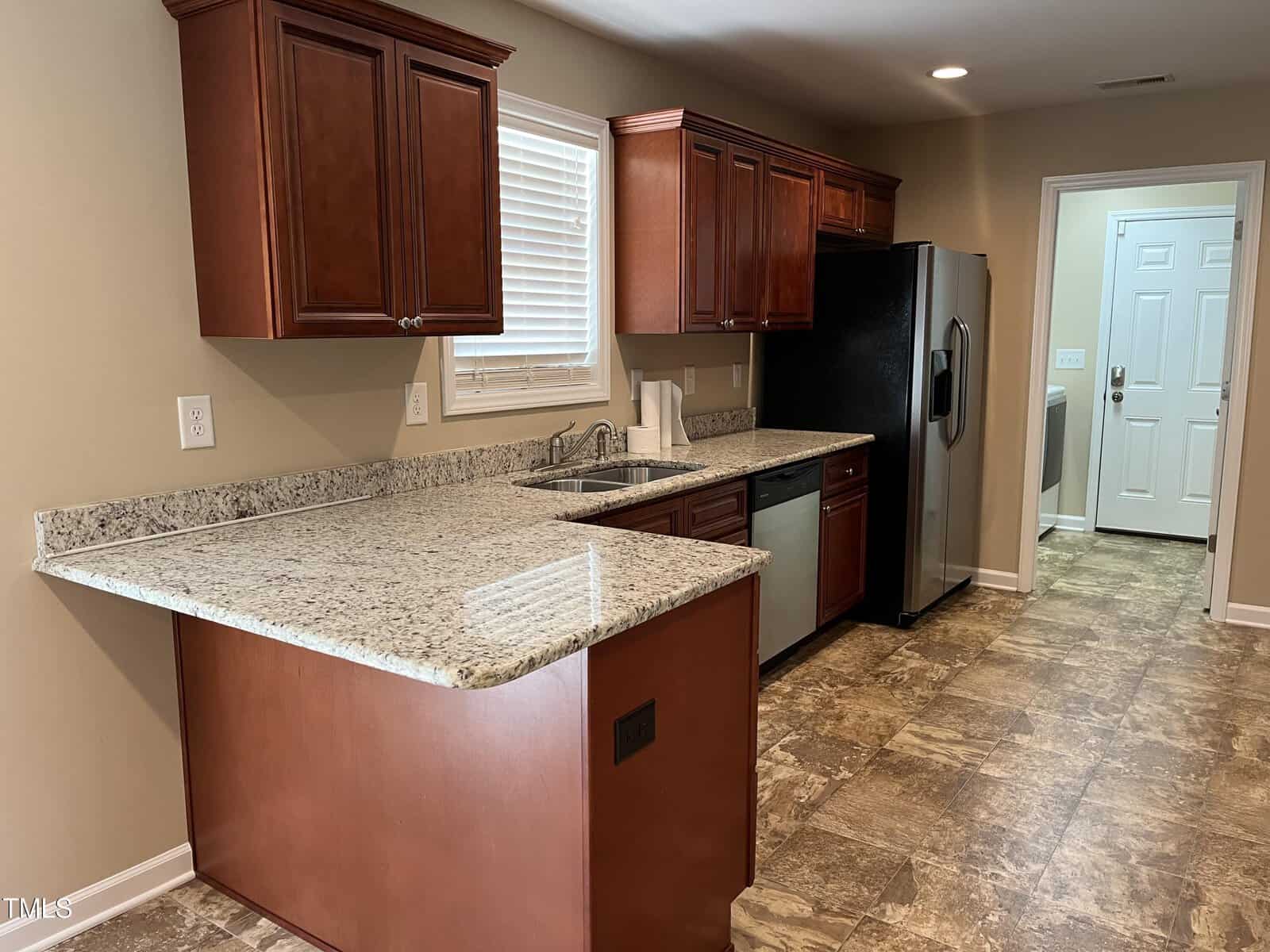 A kitchen with granite countertops, wooden cabinets, stainless steel refrigerator, and a dishwasher. There's a window above the sink and a tile floor. A door in the background leads to another area.