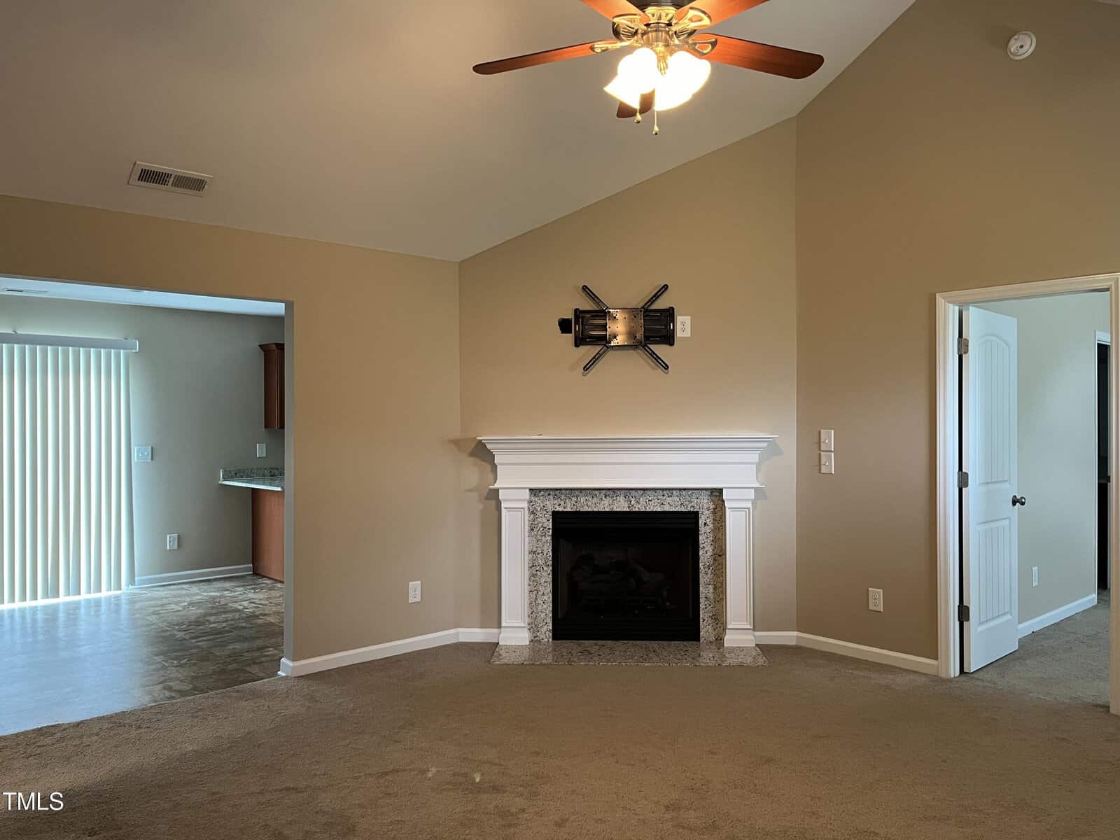 A living room with beige walls and a vaulted ceiling features a ceiling fan with lights. A white fireplace with decorative tiles sits in the center. Open doorways lead to other rooms, and a sliding glass door provides access to an outdoor area.