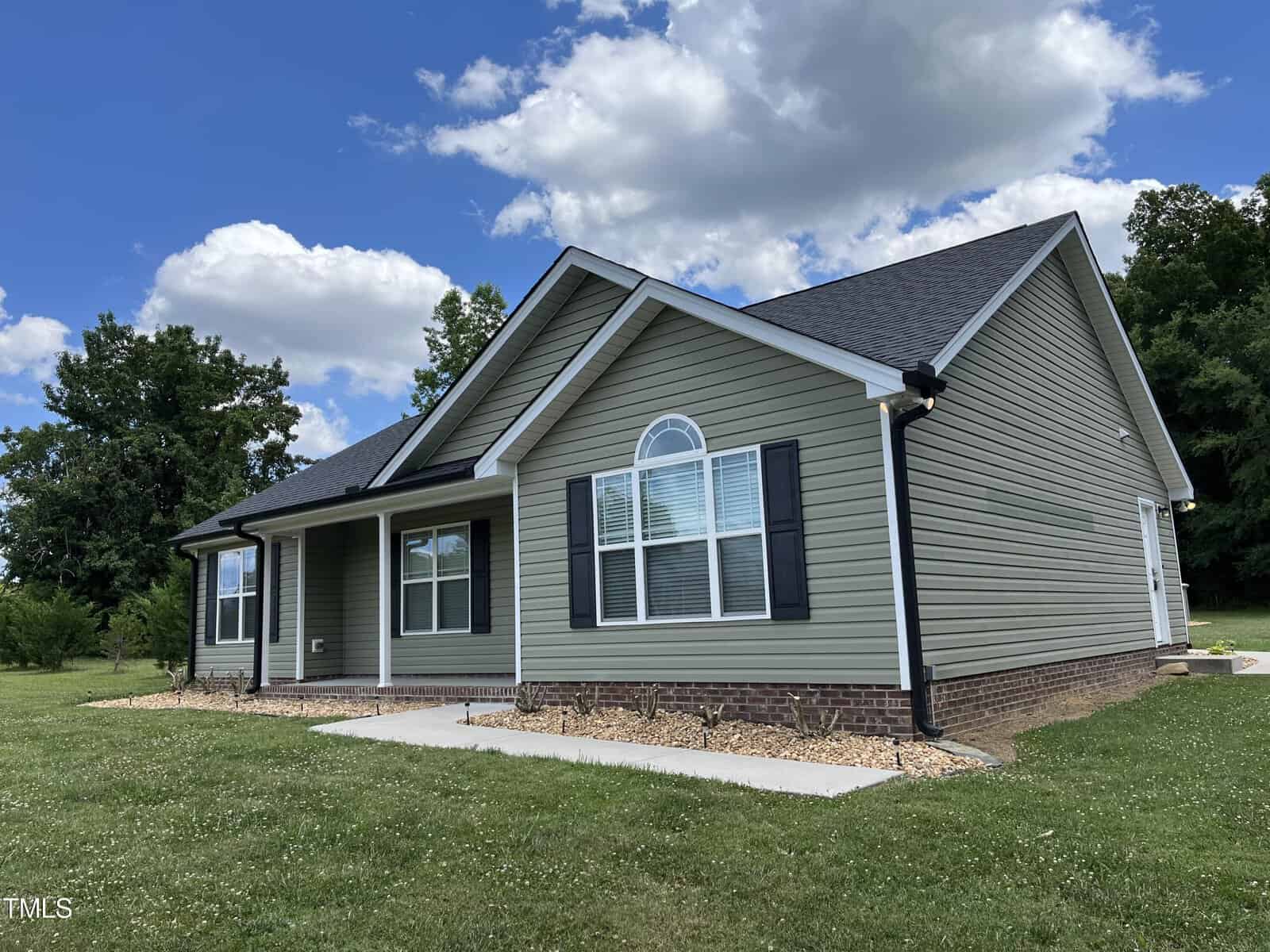 A single-story house with green siding and a dark roof sits in a grassy yard. It features a front porch with white columns and a large arched window. The sky is blue with scattered clouds, and trees are visible in the background.