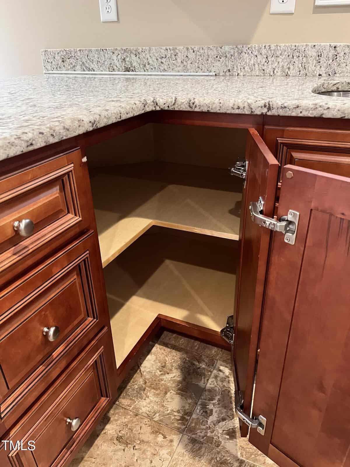 Open kitchen cabinet with brown wood finish and three visible shelves. Adjacent countertop is light-colored granite. Surrounding flooring is brown tile. Cabinet doors are open, revealing empty storage space.