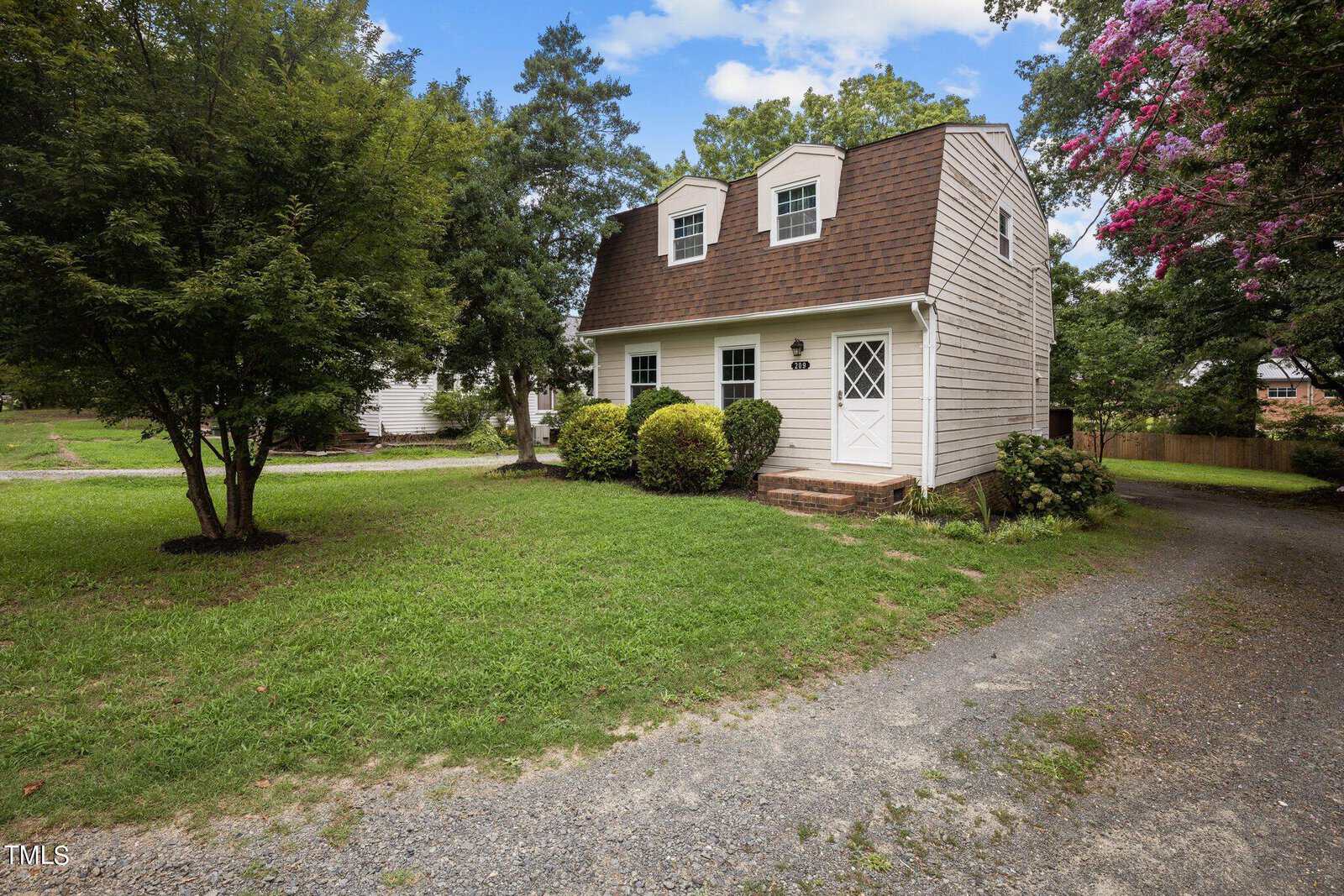Nestled among trees and shrubs, this charming two-story house on 209 Park Avenue in Creedmoor features a gambrel roof and white siding. A gravel driveway leads to a grassy yard, where a small path welcomes you to the elegant white door.