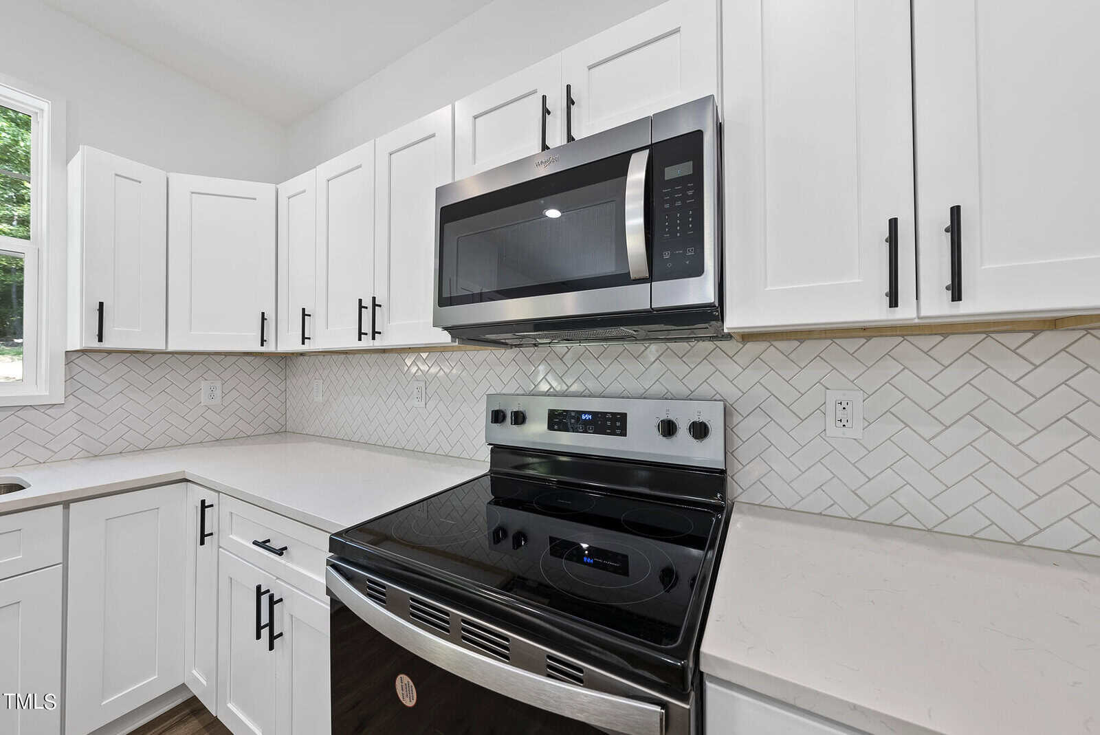 Modern kitchen with white cabinets, stainless steel stove and microwave, white countertops, and a herringbone-patterned tile backsplash. Natural light comes in through a window above the sink.
