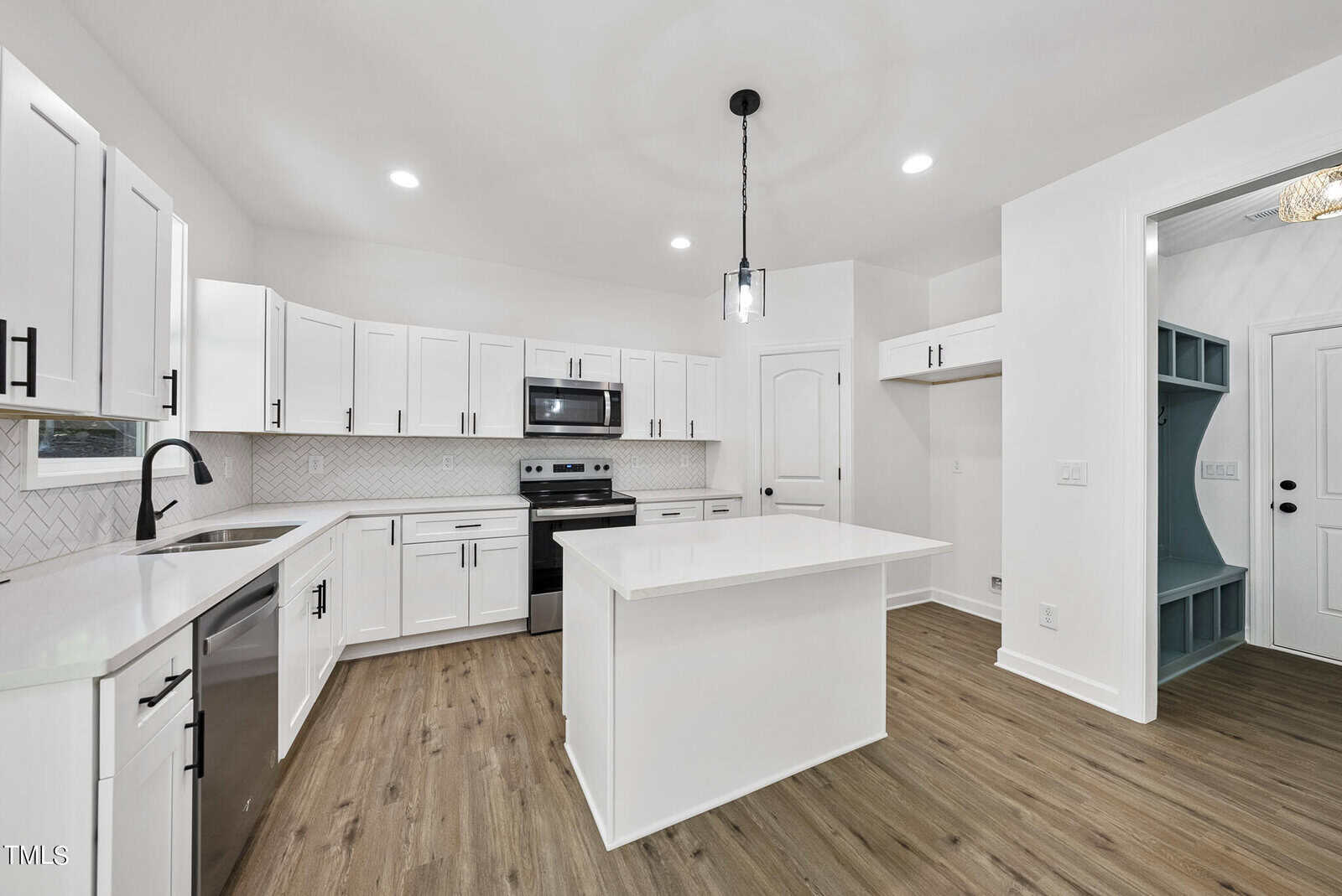 Modern kitchen with white cabinets, stainless steel appliances, center island, light wood flooring, a white backsplash, and recessed lighting. A door opens to a mudroom area with built-in storage and a bench.
