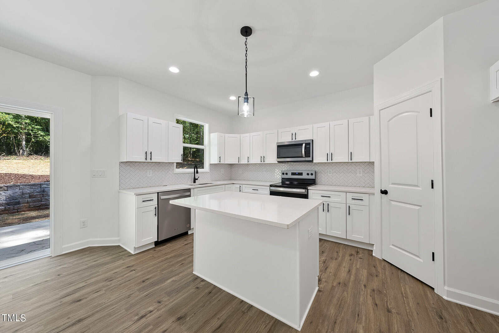 Modern kitchen with white cabinets, stainless steel appliances, a central island, wood flooring, and bright lighting. Window views greenery outside, and a glass pendant light hangs above the island.