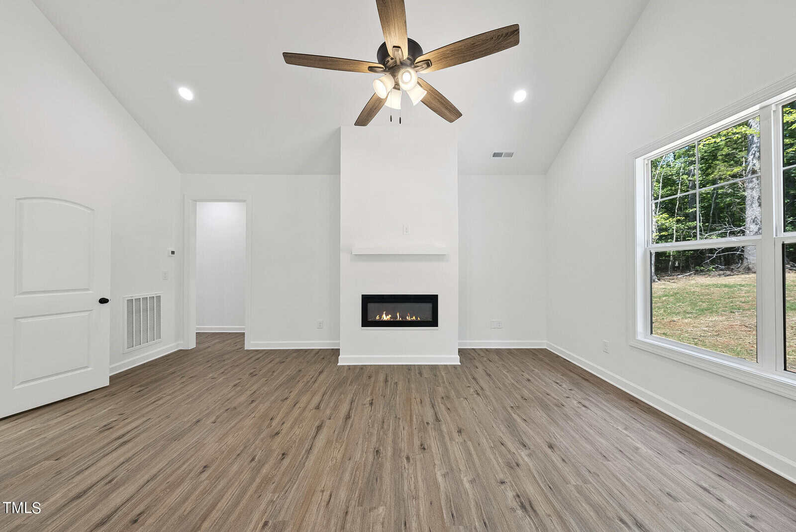 A bright, empty living room with white walls, a modern fireplace, wood floors, a ceiling fan with lights, and a large window showing a view of greenery outside.