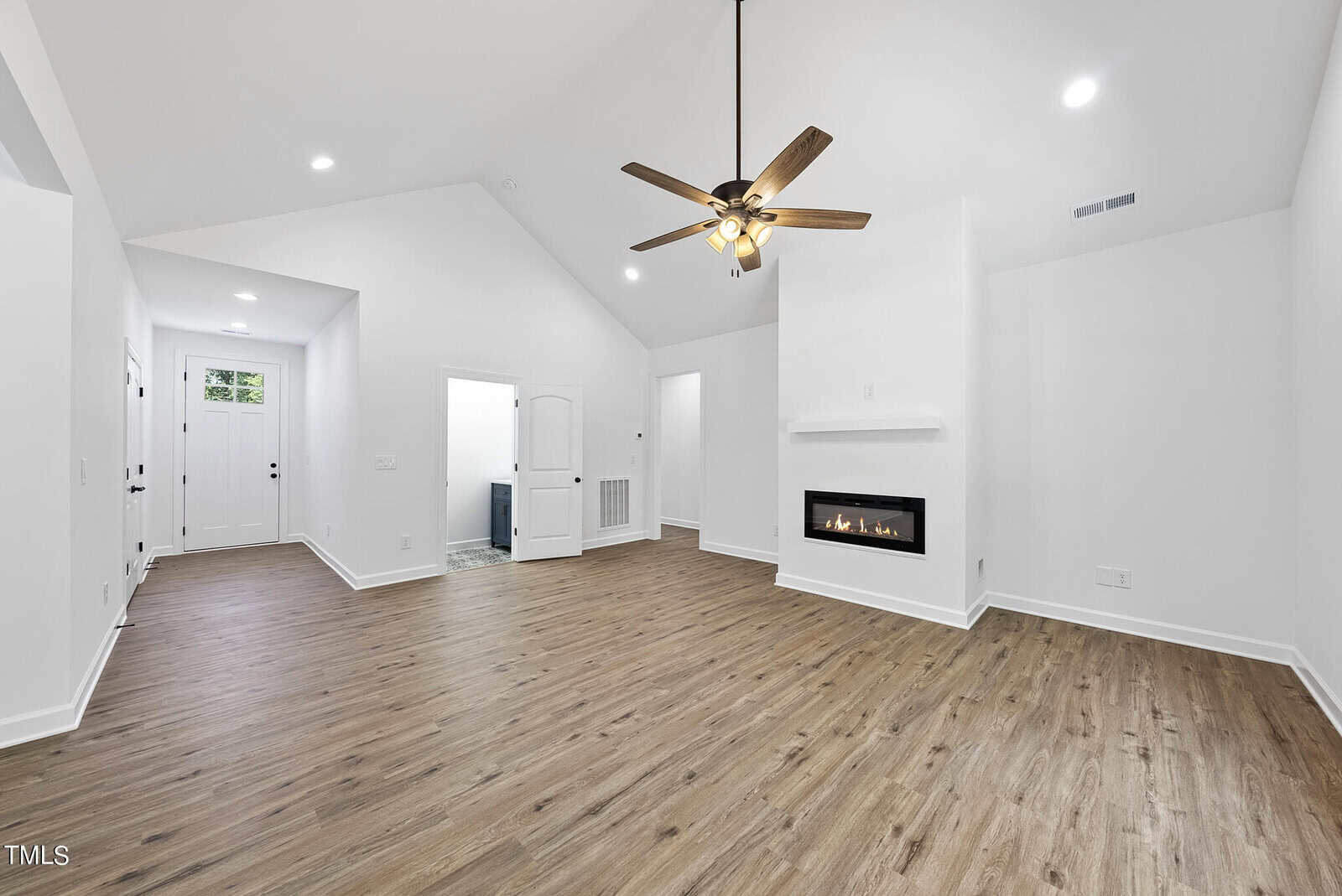 Spacious living room with vaulted ceiling, wood-style flooring, ceiling fan, modern fireplace, and white walls. Entryway and open doorway visible in the background.