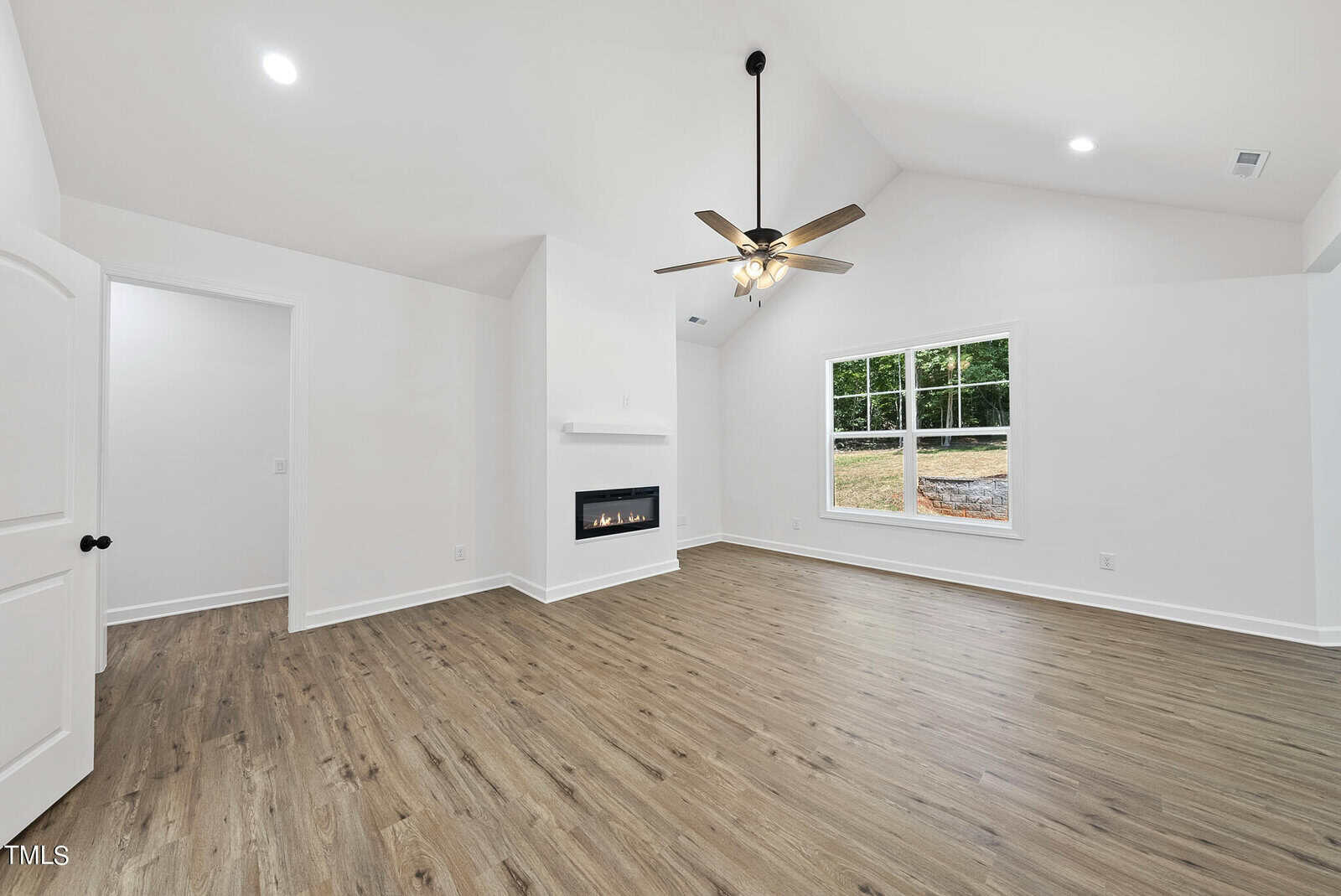 Bright, empty living room with wood-style floors, white walls, a modern fireplace, large window showing a yard, and a black ceiling fan with lights. A white door opens to another room.