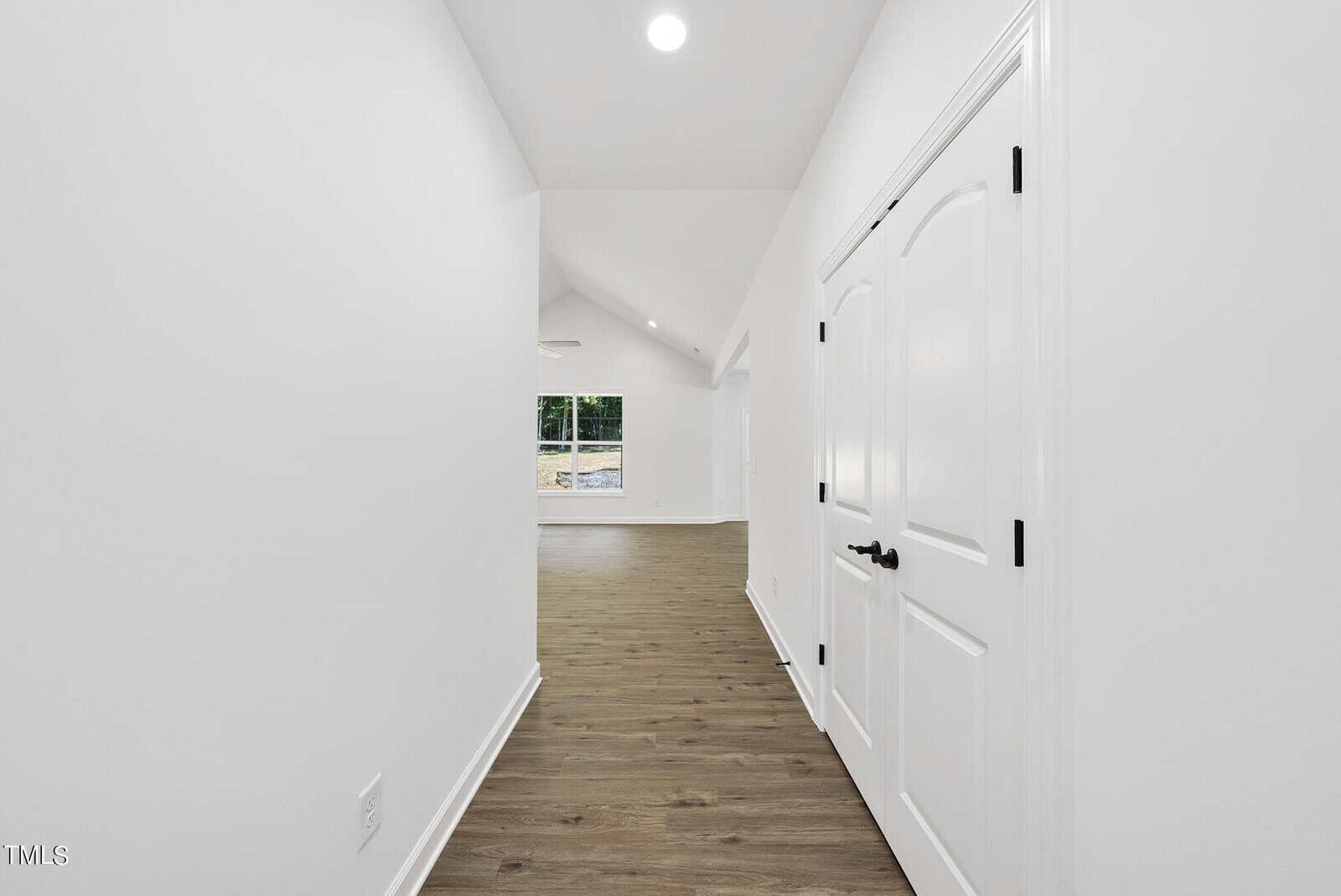 Bright hallway with white walls, vaulted ceiling, wood flooring, and double white doors on the right. The hallway leads to a spacious, well-lit room with large windows at the end.