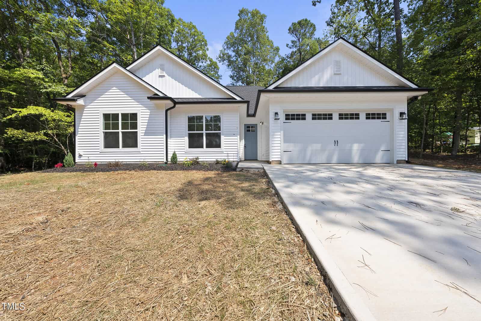 A modern single-story white house with black trim, attached two-car garage, concrete driveway, and minimal landscaping, surrounded by tall green trees under a clear blue sky.