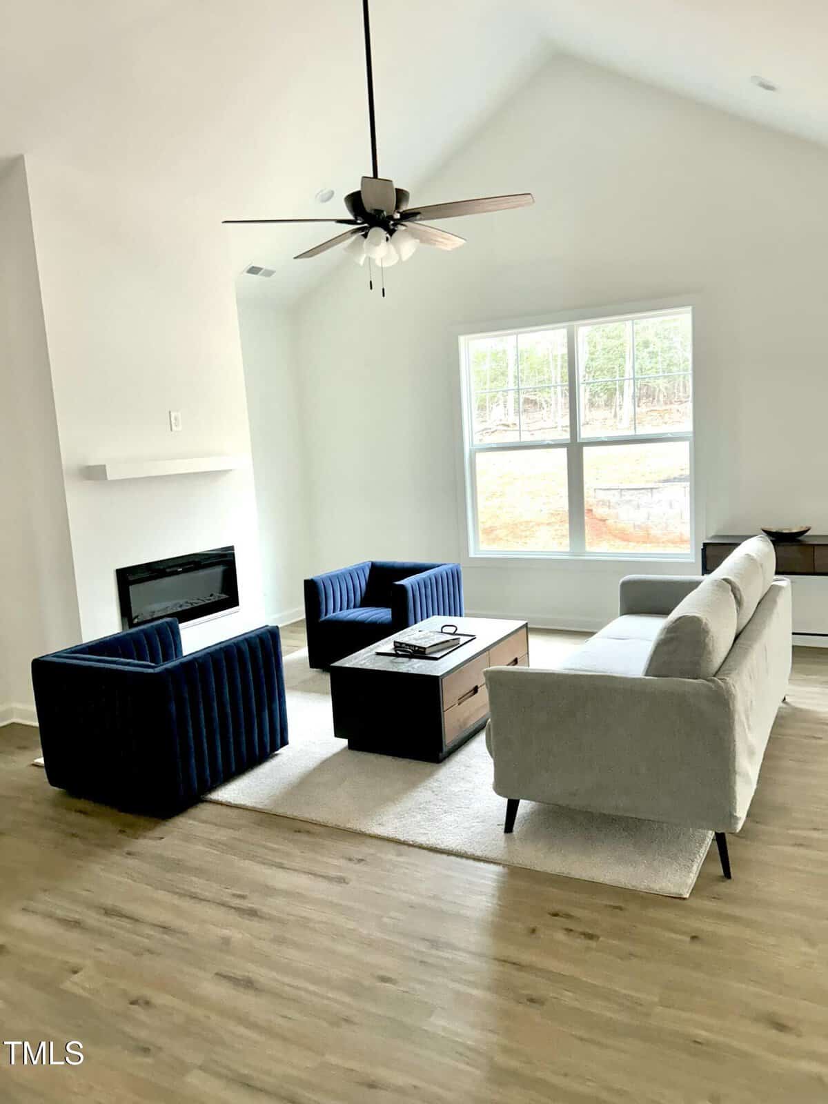 A modern living room with light wood floors, a gray sofa, two dark blue armchairs, a wooden coffee table, a fireplace, and a large window letting in natural light. A ceiling fan hangs from the vaulted ceiling.