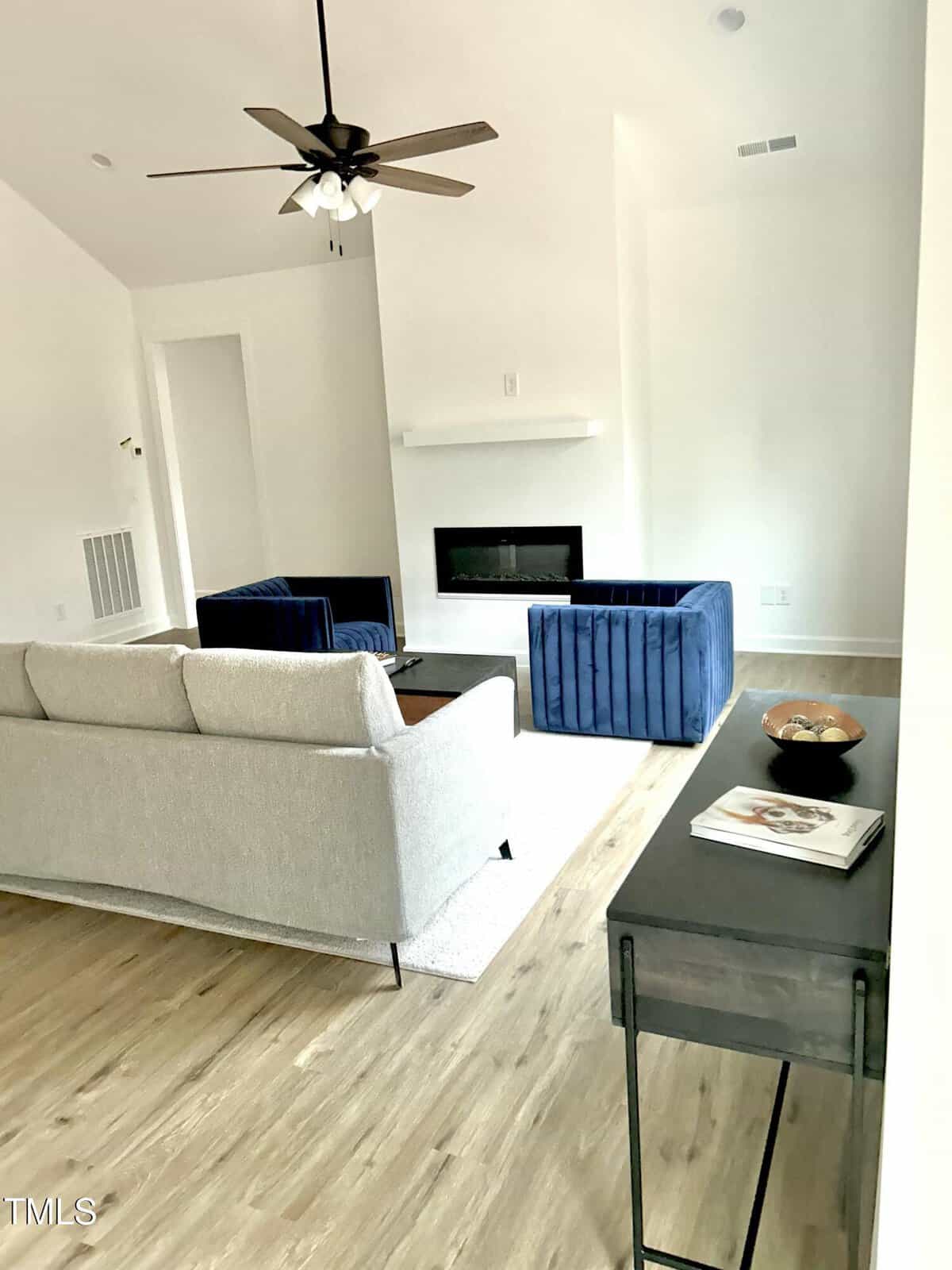 Modern living room with light wood flooring, a beige sofa, two blue velvet armchairs, a black coffee table, and a wall-mounted fireplace. A ceiling fan hangs above, and a black console table sits against the wall.