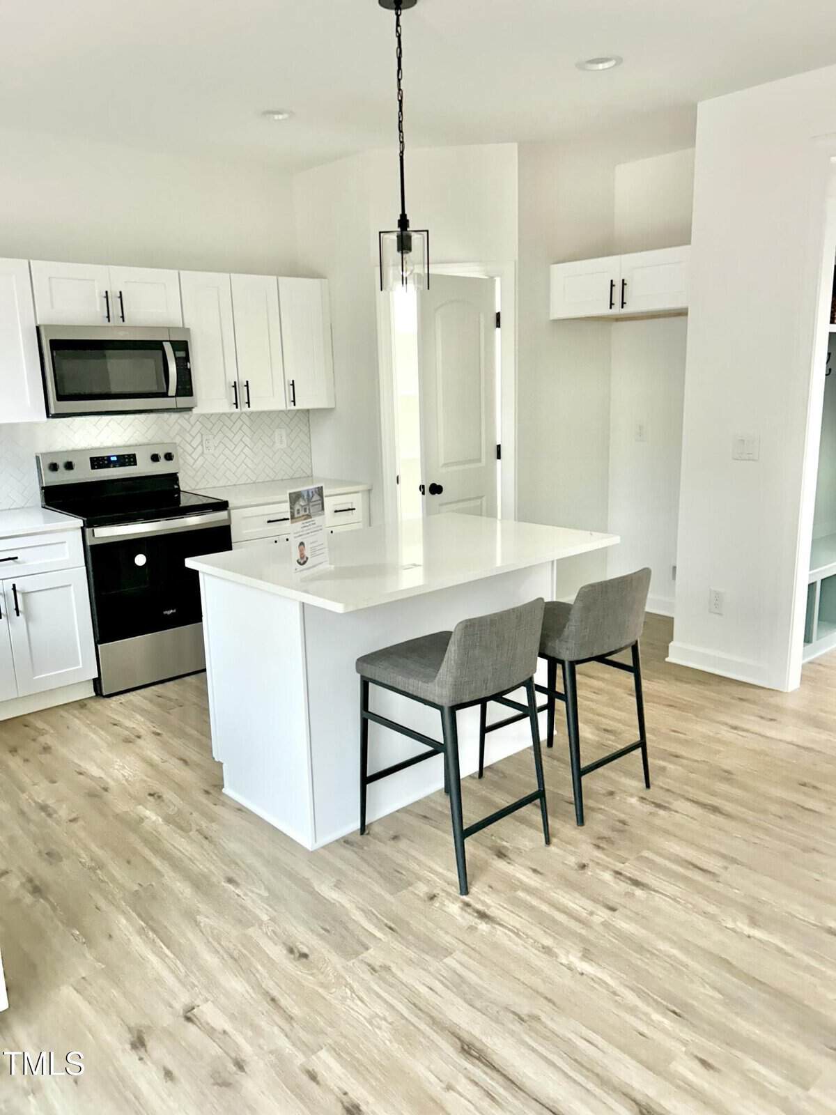 Bright modern kitchen with white cabinets, stainless steel appliances, and a white island with two gray upholstered barstools on light wood flooring. A black pendant light hangs above the island.