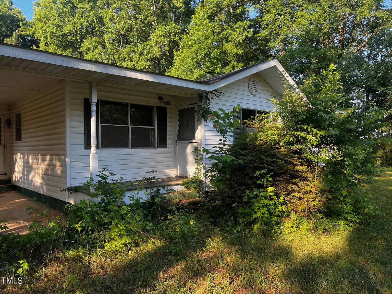 A small white house with a covered porch is nestled among dense greenery at 4018 Eaton Road in Kittrell, with trees and shrubs partially obscuring the front. The house has a pitched roof, large windows, and a door on the front porch. Sunlight filters through the trees, casting shadows on the house.