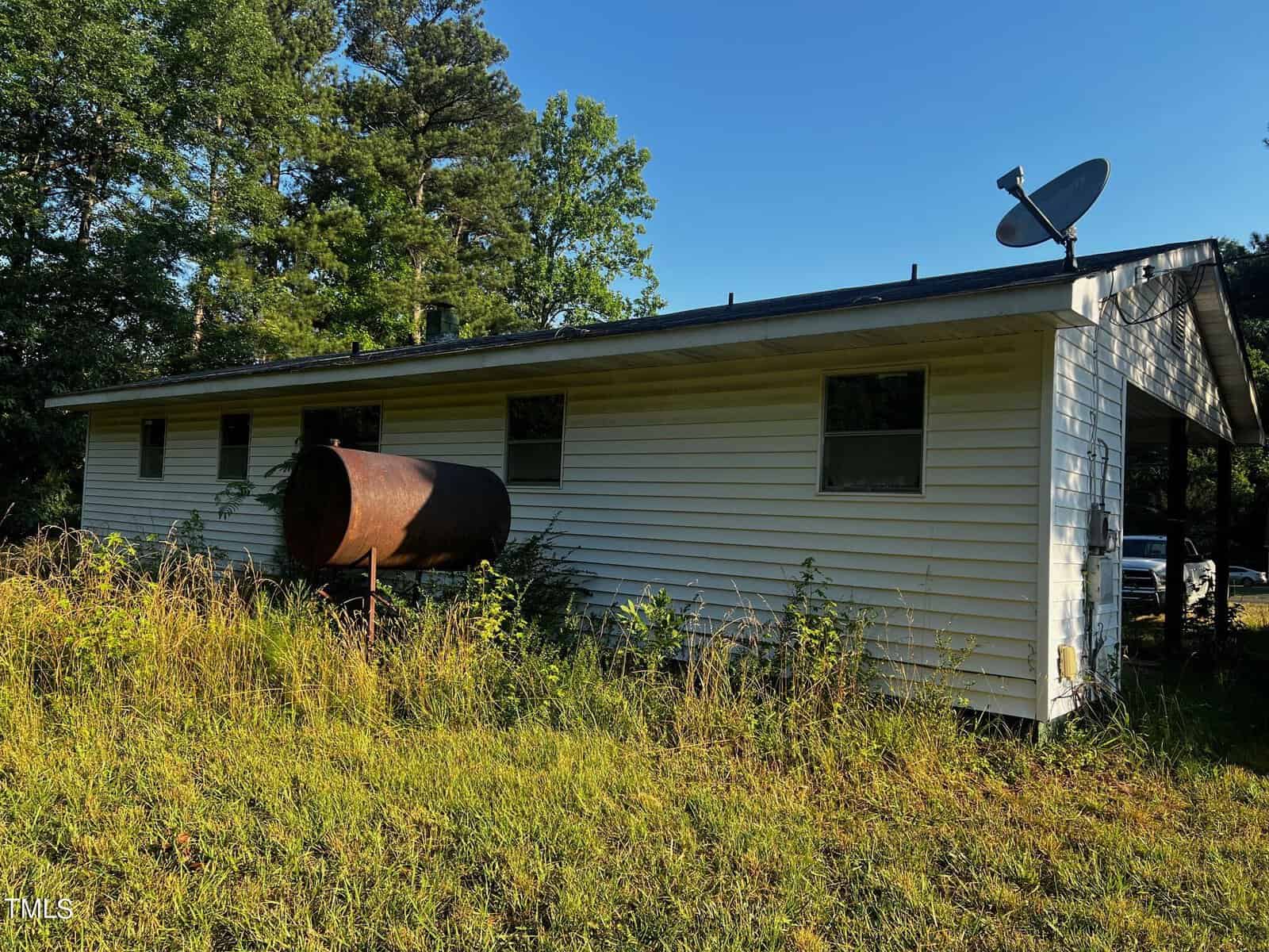 The image shows the side of a small, white, single-story house with a black roof, surrounded by tall grass and trees. An old, rusty oil tank is positioned next to the house. A satellite dish is mounted on the roof, and there are several windows along the side.