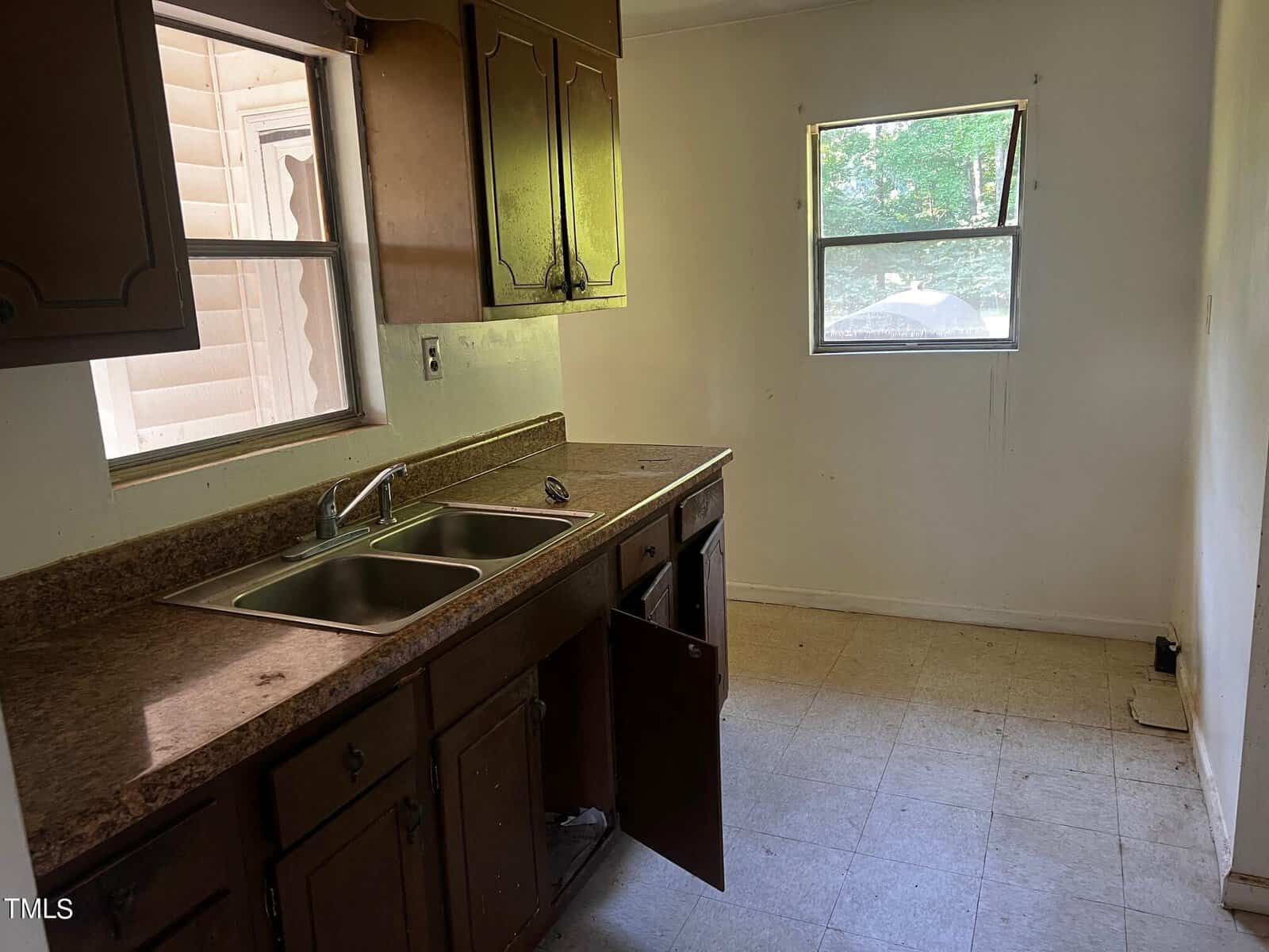 A small kitchen with dark cabinetry, a double stainless steel sink, and brown countertops. One cabinet door is ajar, and there's a window above the sink and another window on the adjacent wall. The floor is covered in light-colored tiles.