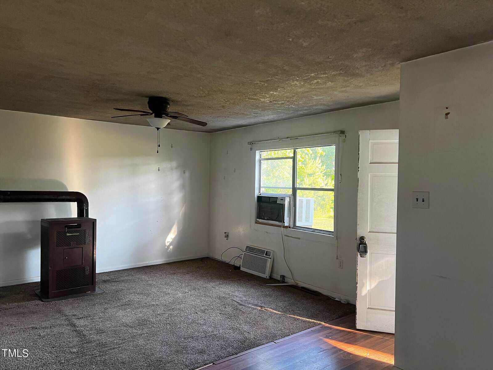 A room with a brown carpet and a white wall. There is an air conditioning unit in a window beside a door. A ceiling fan is mounted on the ceiling. A wood stove is in the corner of the room, and part of the floor is wooden. Natural light is coming in.