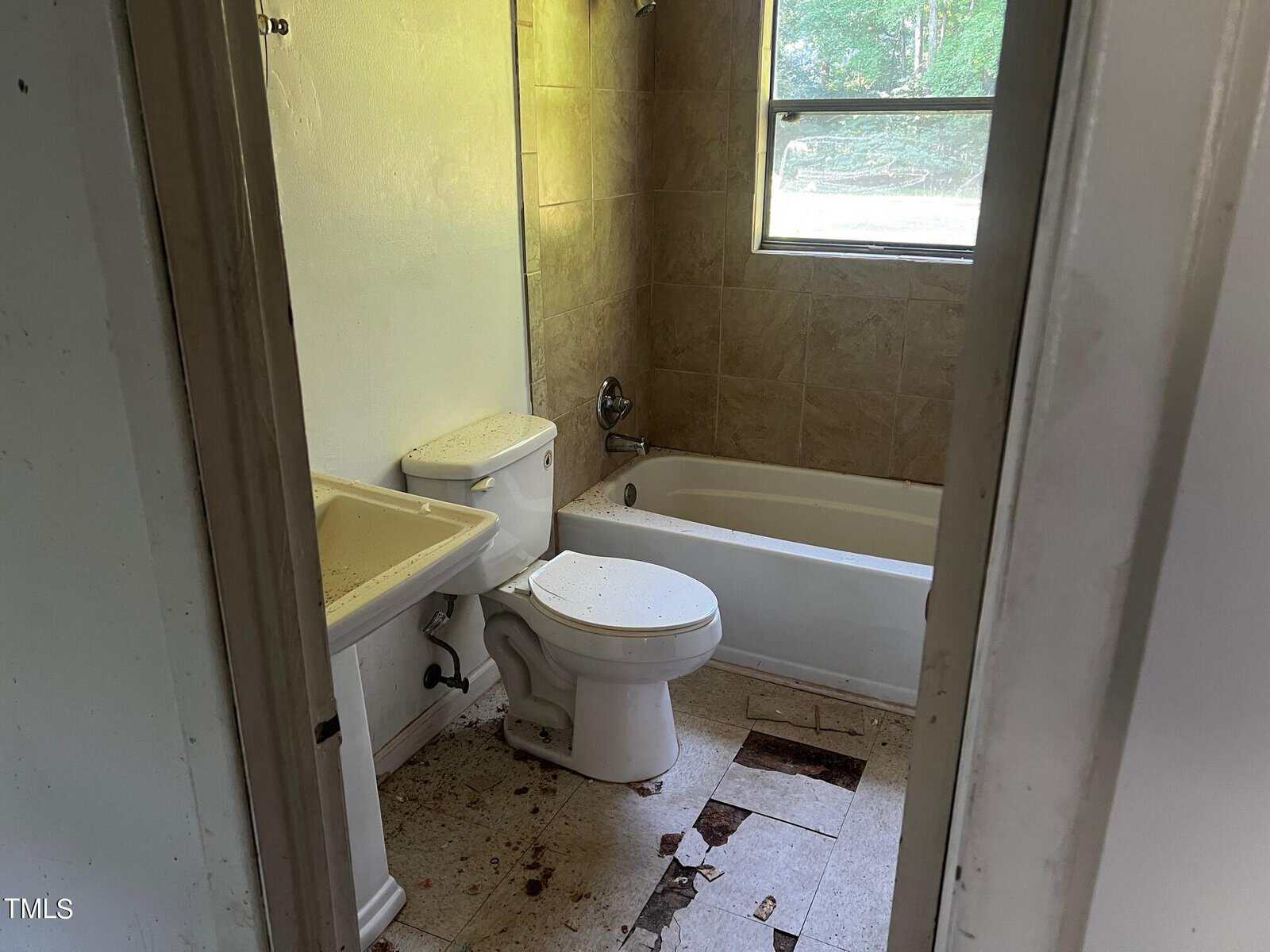 A small bathroom with a white sink, toilet, and bathtub. The wall above the bathtub is tiled. The floor has several broken and missing tiles, and there are visible dirt and stains. A window above the bathtub lets in natural light from outside.