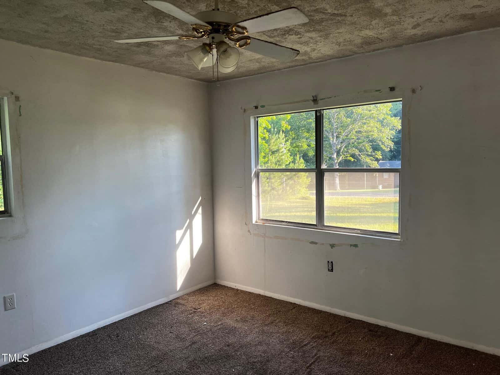 A small, empty room with a ceiling fan and two windows. The walls are bare and painted white. The carpet is brown and appears worn. Sunlight is streaming in through the windows, illuminating part of the room. The condition of the room suggests it may need some renovation.