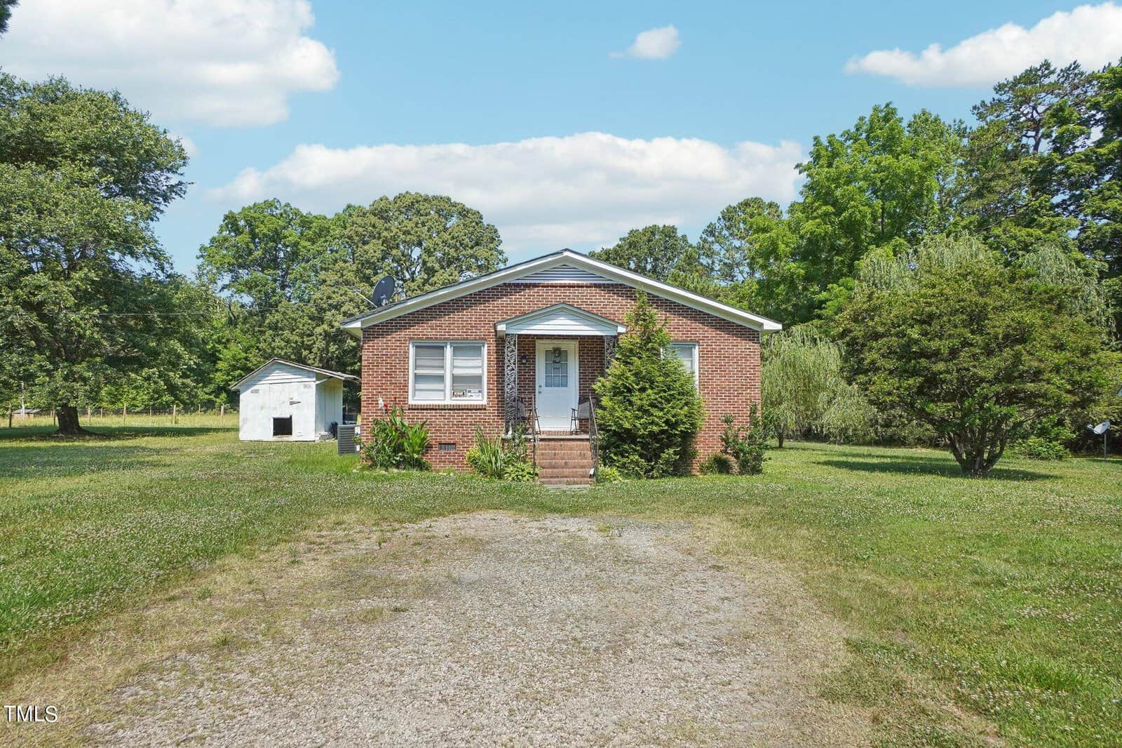 A small brick house with white trim is situated in the center of the image. The house is surrounded by a large grassy yard with a gravel path leading to the front steps. A white shed is visible to the left, and trees border the yard in the background. The sky is clear with some clouds.