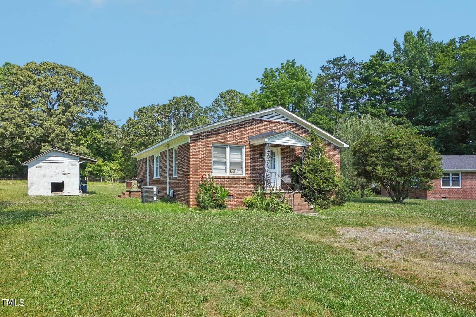 A small, single-story red brick house with a gabled roof and white trim sits in a grassy yard. It features a few steps leading to the front door and is surrounded by greenery and trees. A small detached building is seen in the background. The sky is clear and blue.