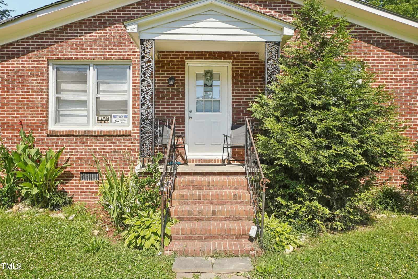 A brick house with a small front porch featuring decorative black metal railings and two chairs. A white door is at the top of several brick steps leading to the porch. There are landscaped plants and grass in the front yard, along with bushes next to the porch.