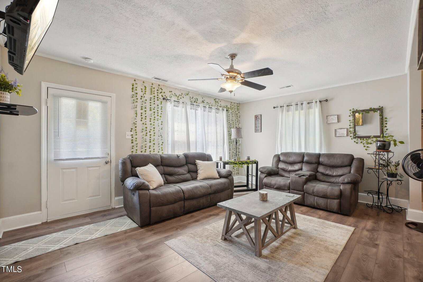 A cozy living room with light grey walls, a ceiling fan, and large windows covered with sheer white curtains. The room features two brown leather recliners, a wooden coffee table, and green vine wall decorations. A small table is placed between the recliners. A door is on the left.
