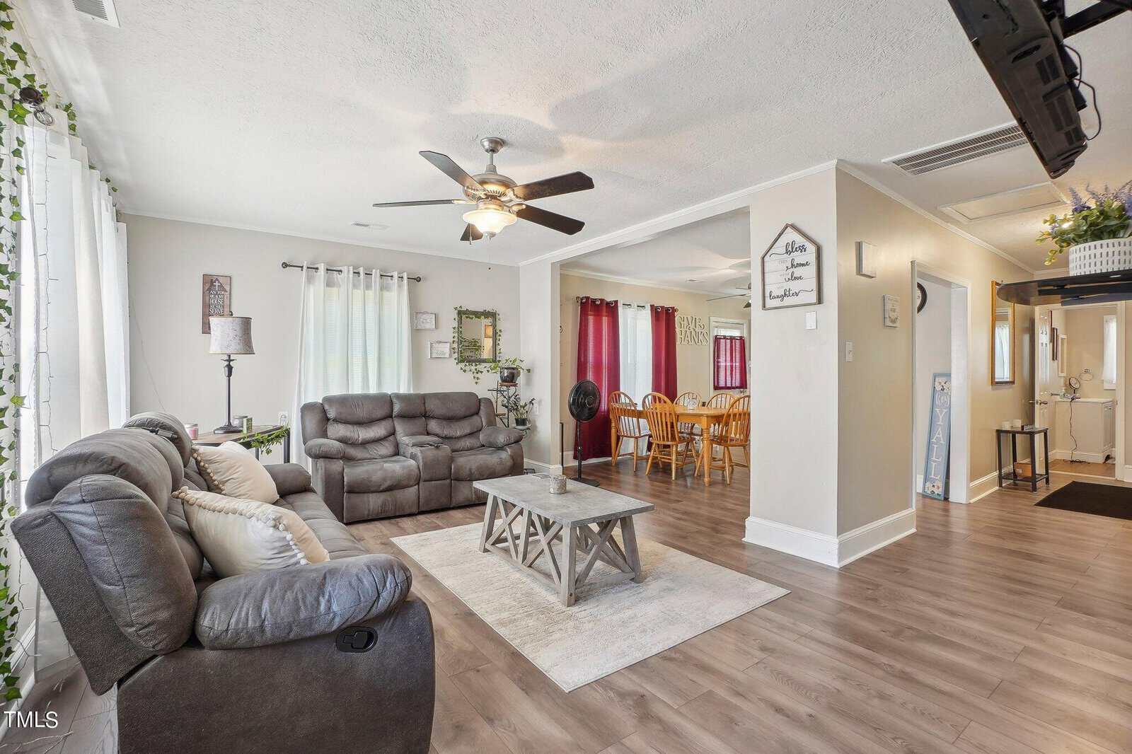 A cozy living room with a brown sectional sofa, recliner, and a wooden coffee table in the center. A ceiling fan hangs from the white ceiling. The room leads into a dining area with a wooden table and chairs. Light streams in from large windows with white curtains.