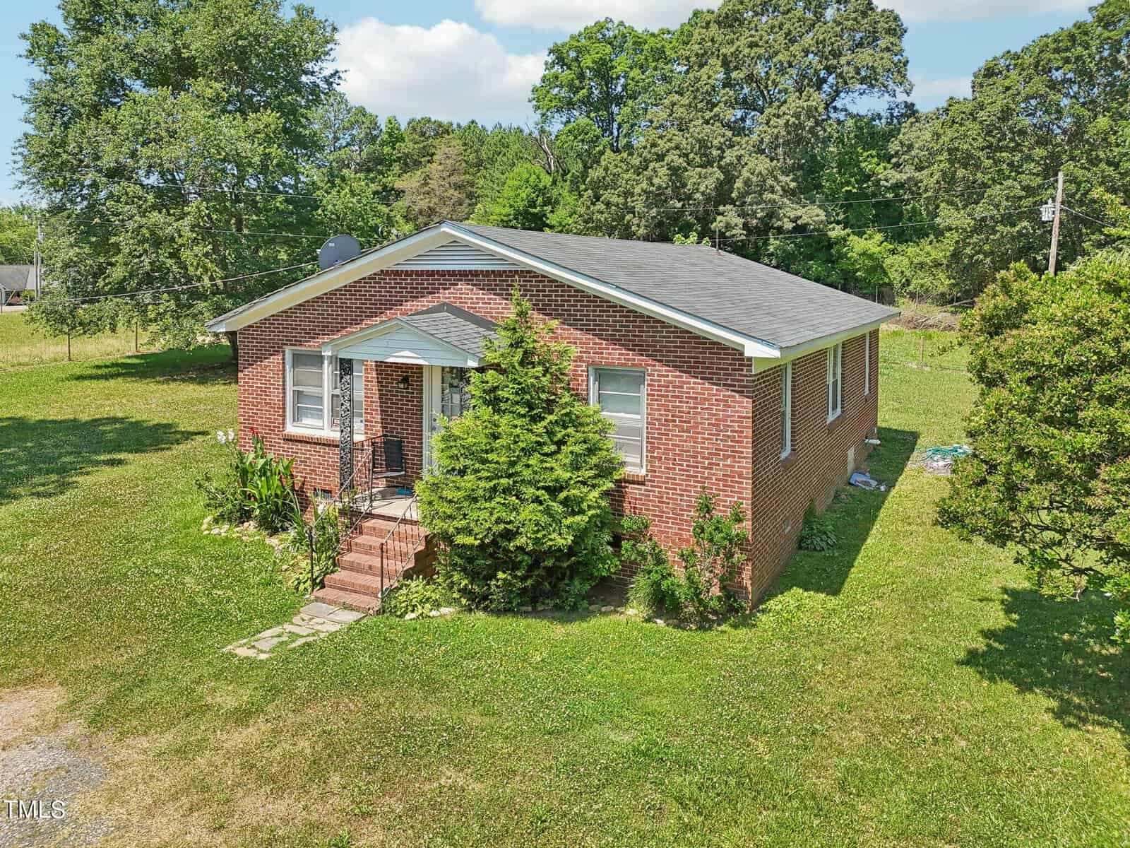 A single-story red-brick house with a gray roof sits on a grassy lawn. There's a small front porch with steps leading up to a white-framed door. Green shrubs and trees surround the house, and a large tree is in the background. The sky is clear with some clouds.