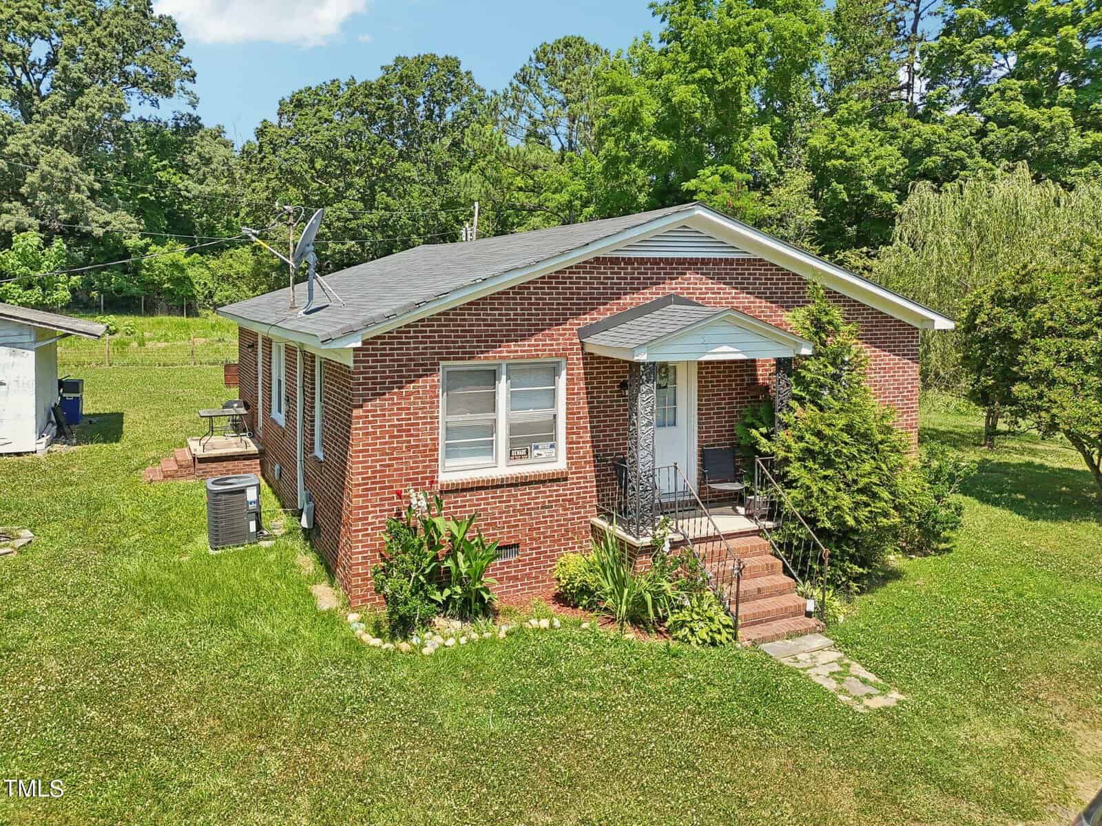 A single-story red brick house with a small front porch and steps. The house is surrounded by a lawn with green trees and shrubs. A satellite dish is mounted on the roof, and there is an air conditioning unit on the side of the house. The sky is clear and sunny.