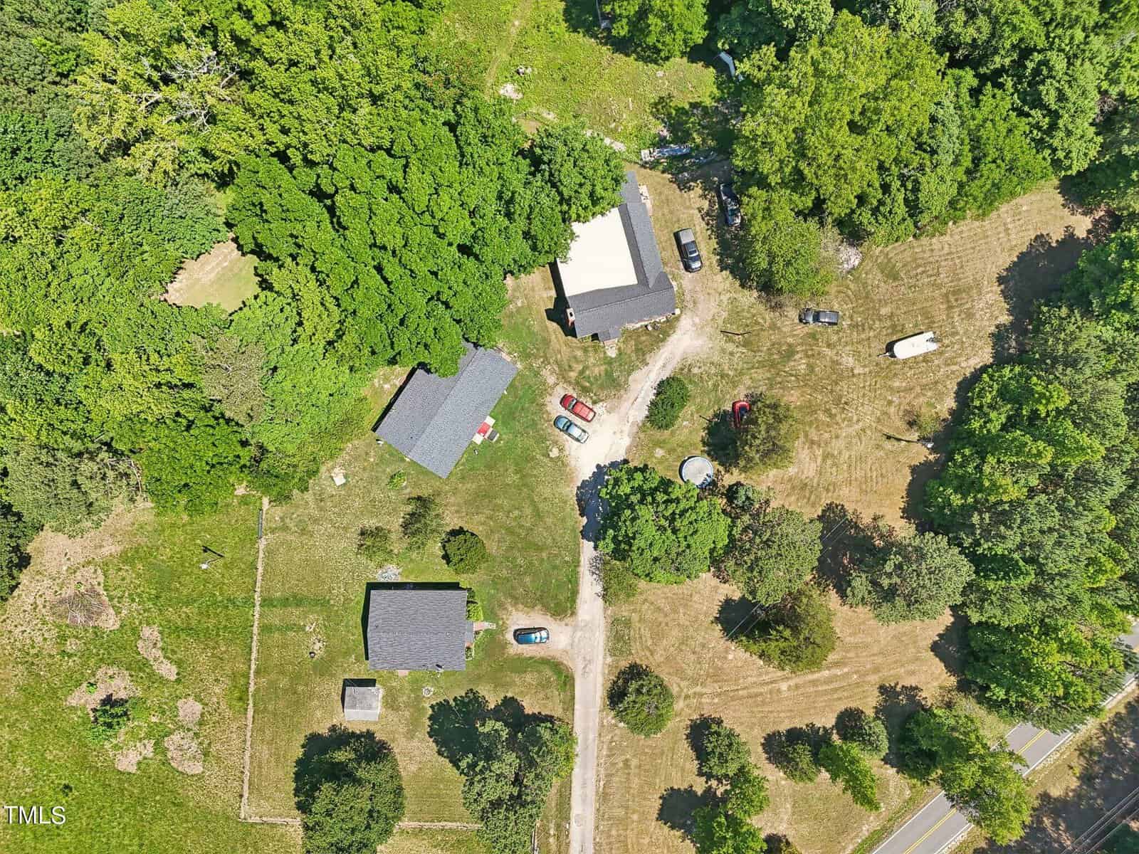 Aerial view of a rural property featuring three buildings surrounded by dense trees and open grassy areas. Several vehicles are parked on the property, and a dirt road winds through the center. The landscape includes a mix of cleared land and wooded areas.