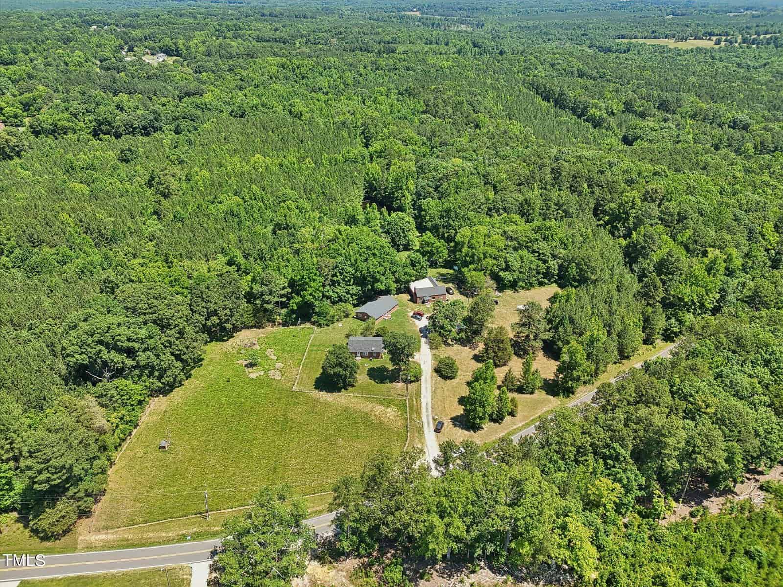 Aerial view of a rural property surrounded by dense forest. The property includes several buildings, open grassy areas, and a driveway that leads to a main road. The lush green landscape extends into the distance, depicting a serene and secluded setting.