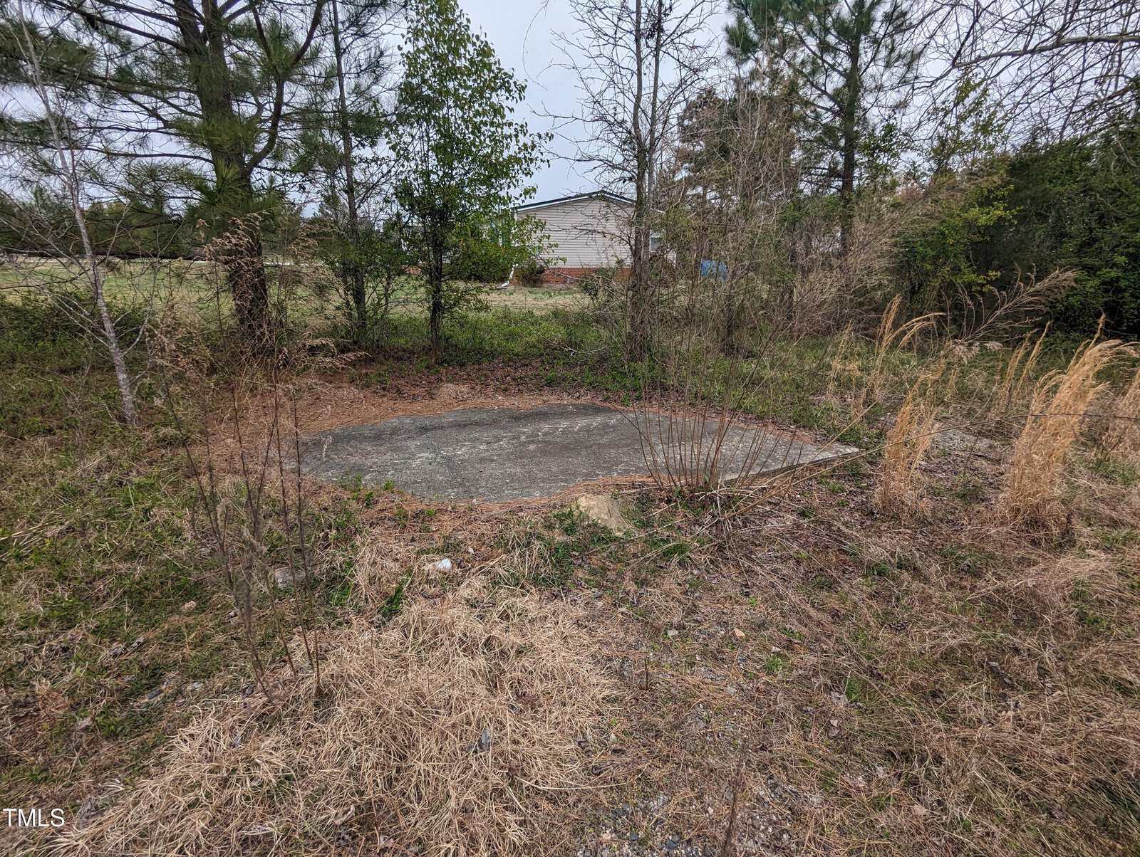 A small, rectangular concrete slab is surrounded by overgrown vegetation, including grass and trees, in a rural area. A large building is visible in the background under a cloudy sky.
