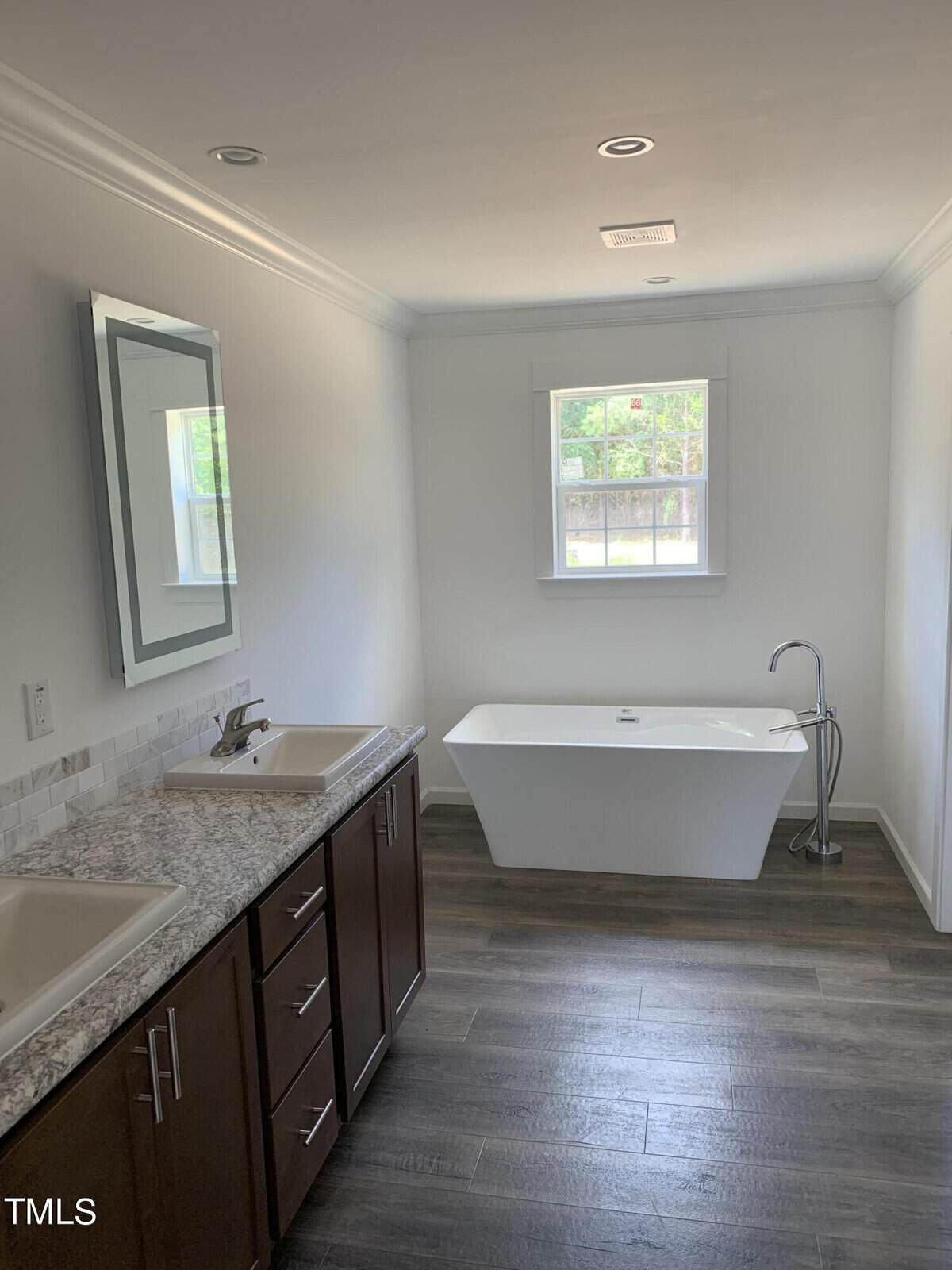 A modern bathroom with a freestanding bathtub next to a window, double vanity with granite countertop and undermount sinks, dark wood cabinets, and wood-look tiled flooring. A large mirror is mounted above one of the sinks. The room is well-lit with natural light.