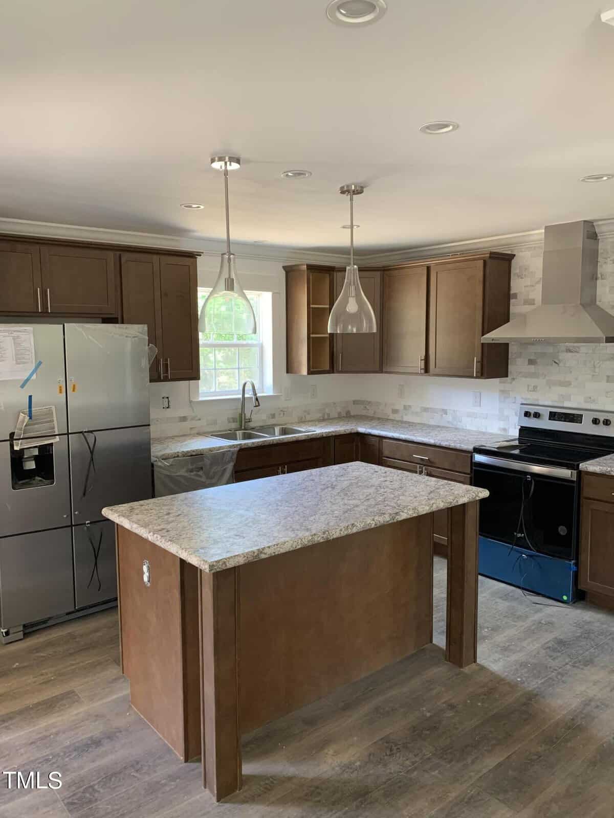 Modern kitchen interior featuring dark wooden cabinetry, stainless steel appliances including a refrigerator and stove, marble countertops, and an island with a marble surface. Two pendant lights hang above the island, and a window provides natural light.