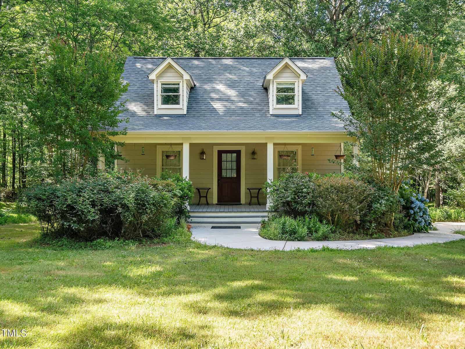 A charming small house at 3637 Sandy Creek Road features a gray roof, two dormer windows, and a cozy covered front porch. Nestled in Franklinton's lush greenery and trees, the home offers a welcoming walkway leading to the entrance, with two chairs ready for relaxation on the porch.