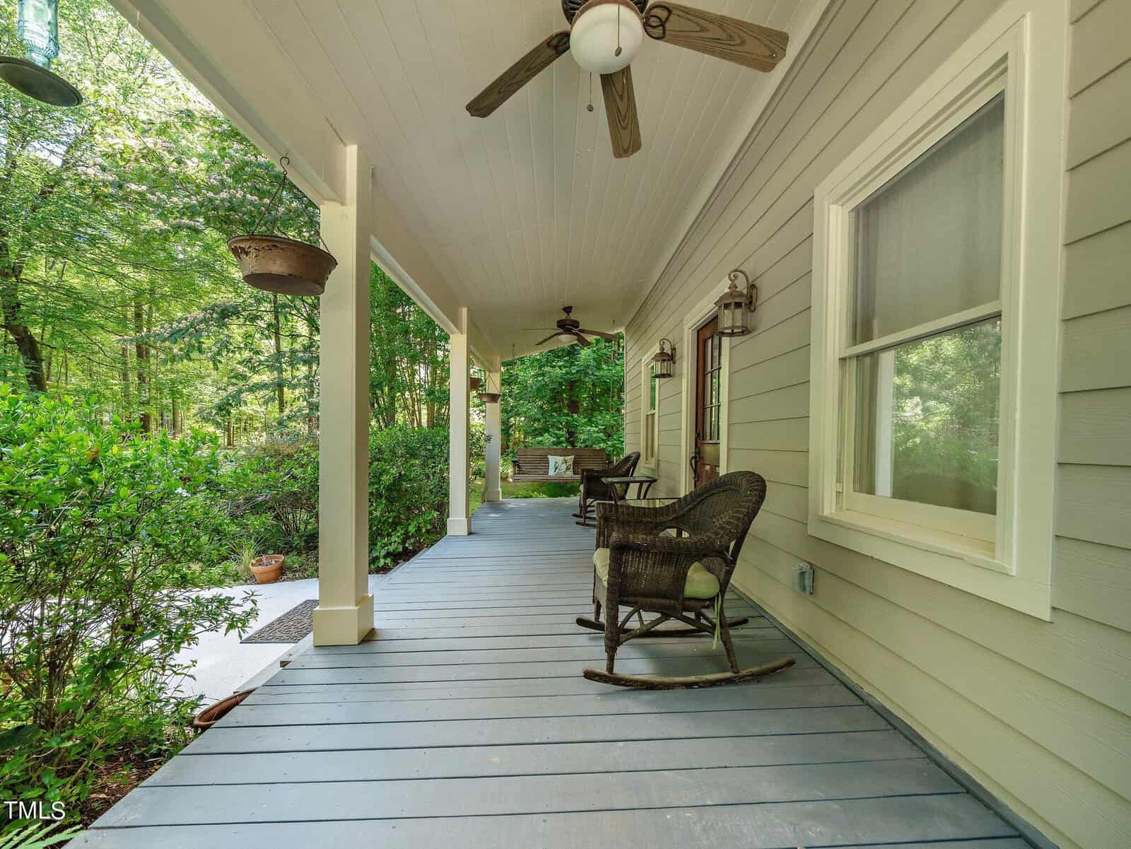A covered porch with two wicker rocking chairs and hanging plants. Ceiling fans are mounted above, and a lush green garden surrounds the area, creating a serene and inviting atmosphere.