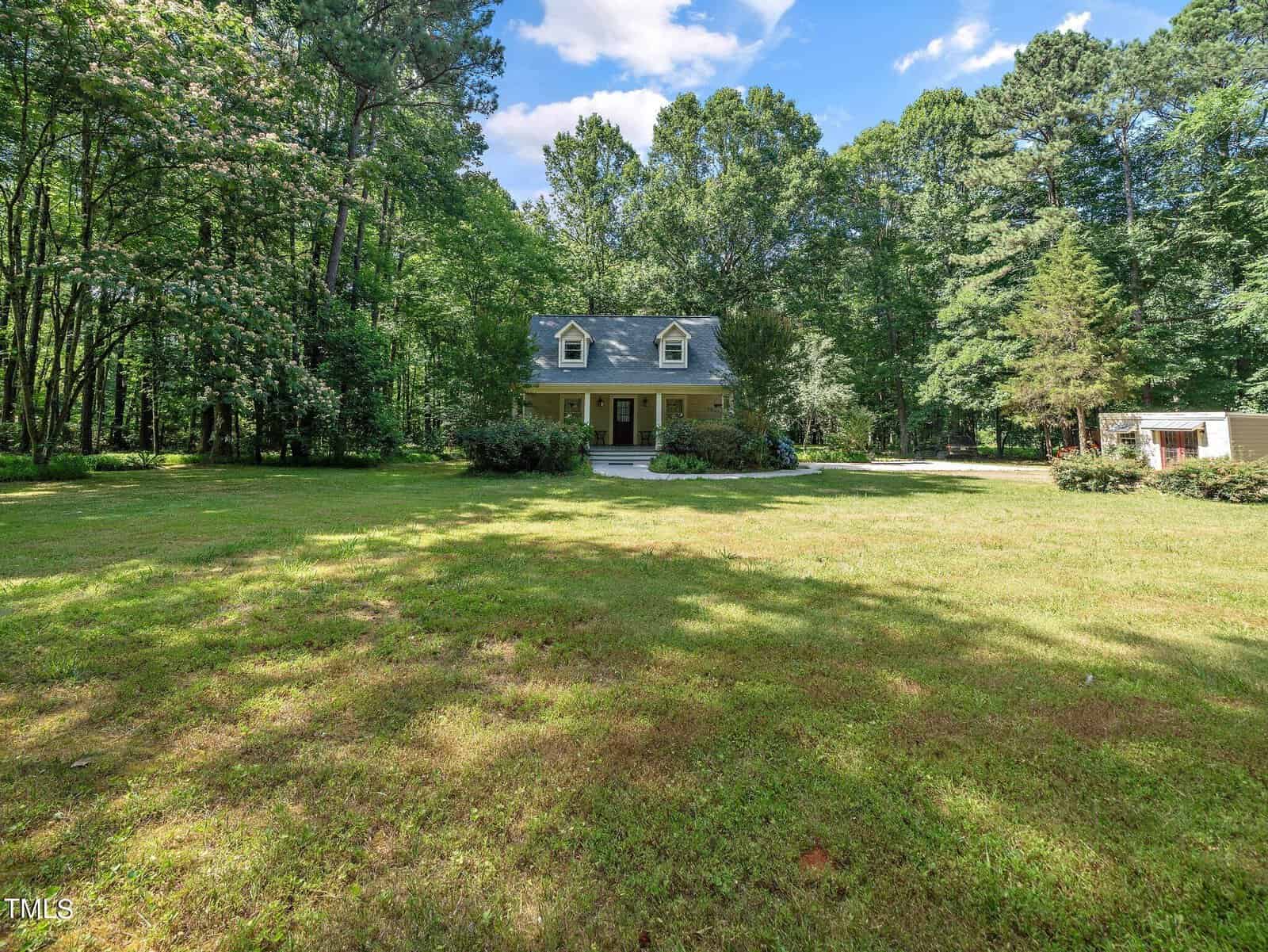 A yellow two-story house with a porch, surrounded by lush green trees and a large grassy yard under a clear blue sky. A small detached structure is on the right.