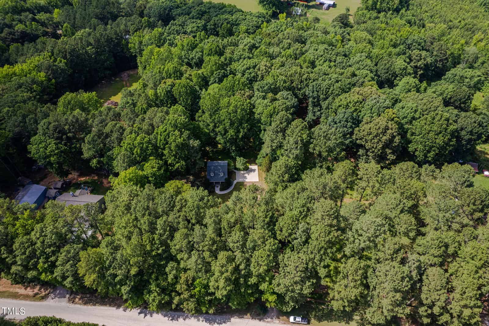 Aerial view of a dense forest with a single-story house partially visible in the center. The house is surrounded by tall green trees, and a winding driveway leads to a gravel road at the bottom of the image. Small clearings are visible nearby.