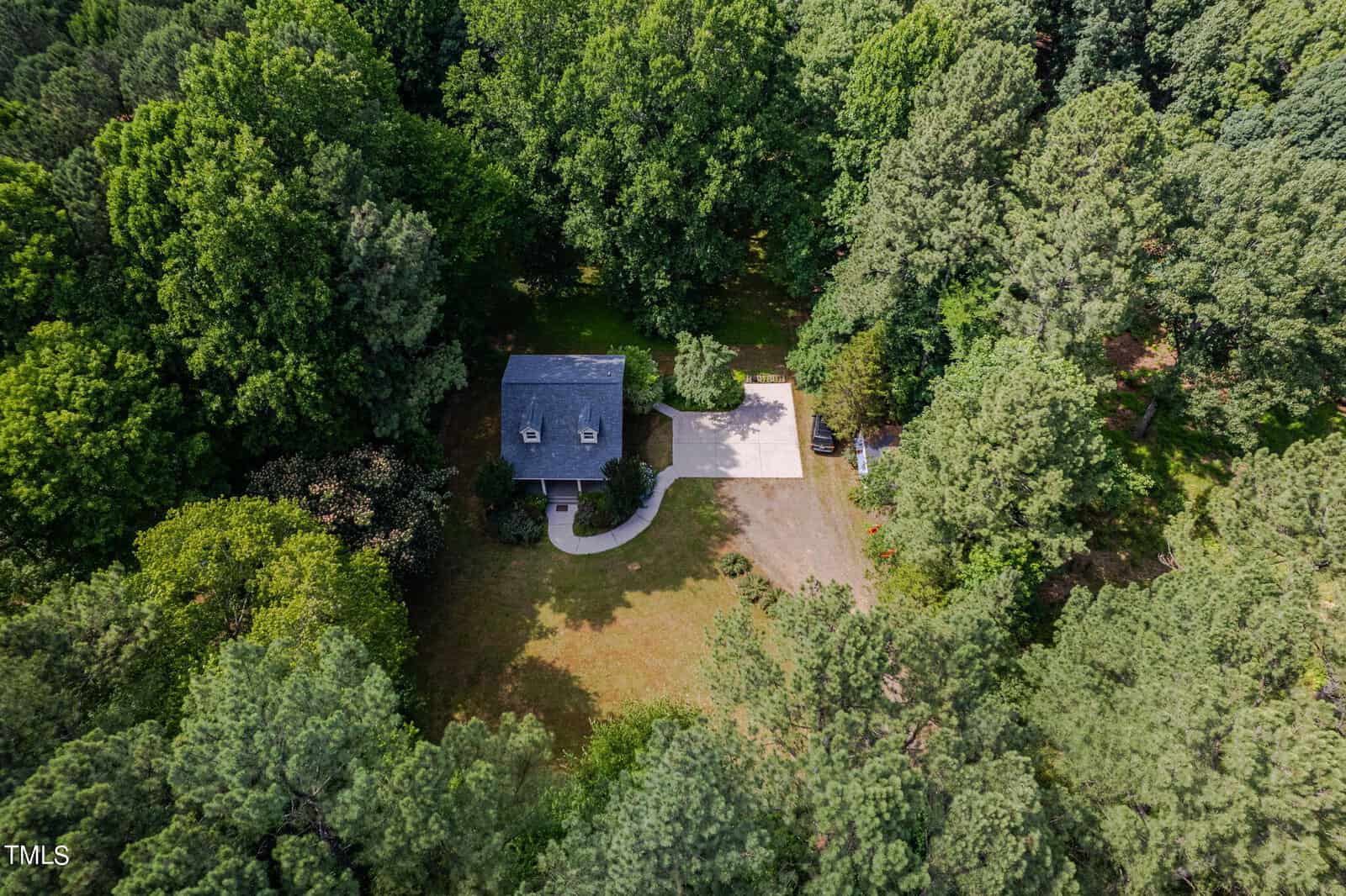 Aerial view of a small house surrounded by dense green trees. The property includes a driveway leading to the house and a large garage or shed nearby. The area appears serene and secluded, with a mix of mature and younger trees.