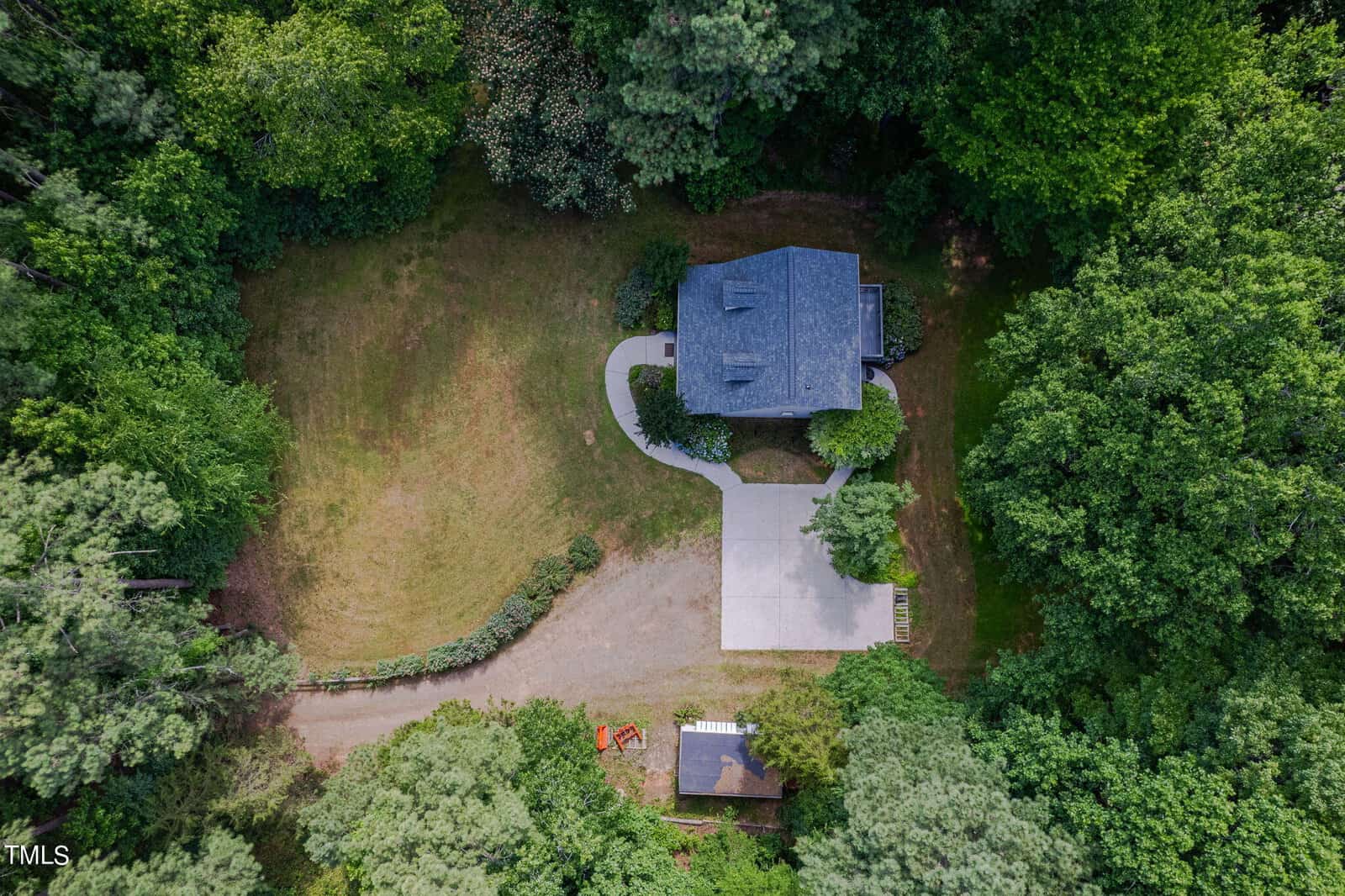 Aerial view of a house surrounded by dense green trees. The property features a large, partially paved yard, a curved driveway, and a smaller structure below. A small garden or vegetated area is visible near the house.