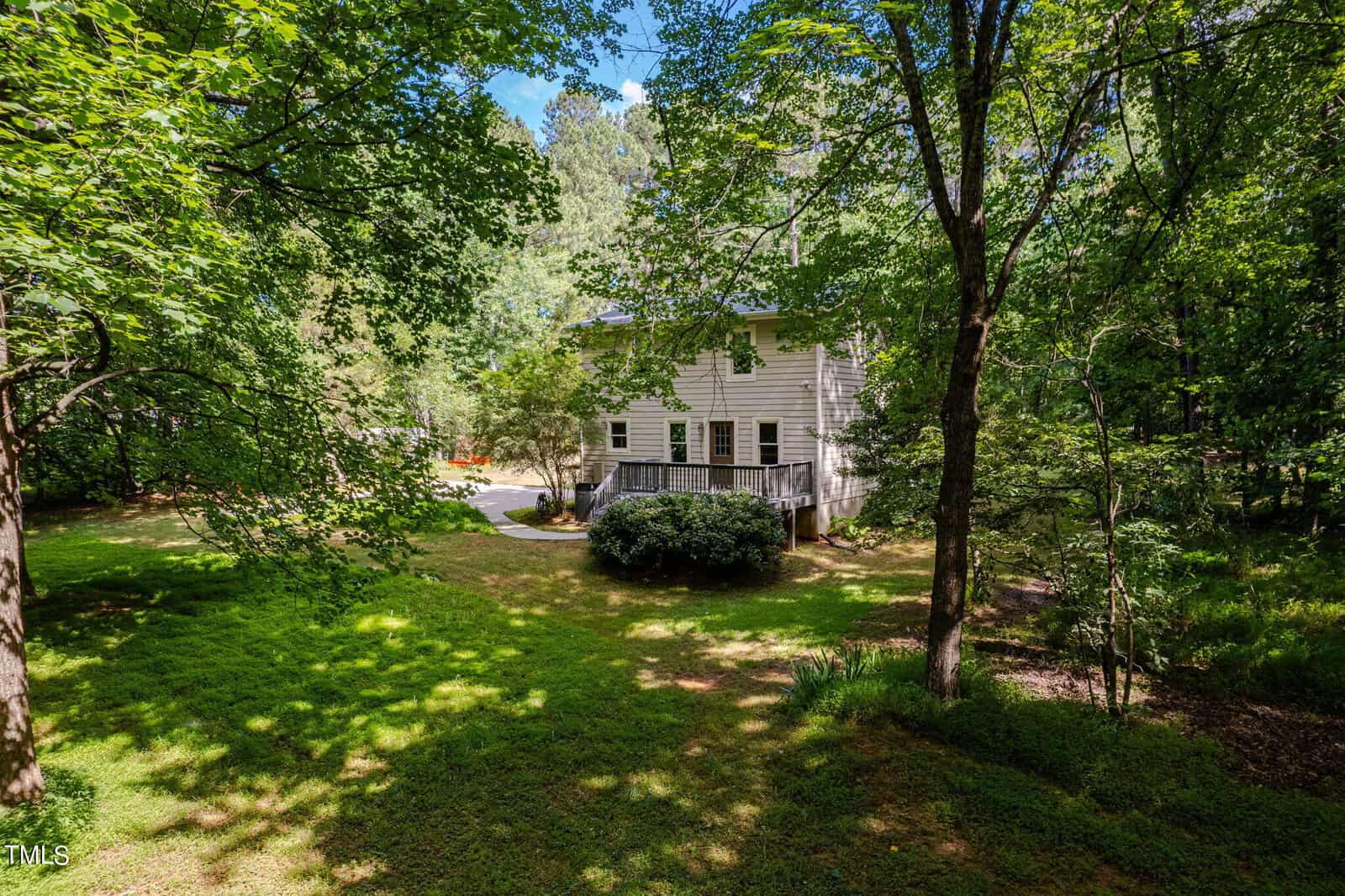 A two-story house with beige siding is nestled among tall, leafy trees. It features large windows and a deck. The surrounding yard is lush with grass and shaded by greenery, creating a serene, forest-like setting.