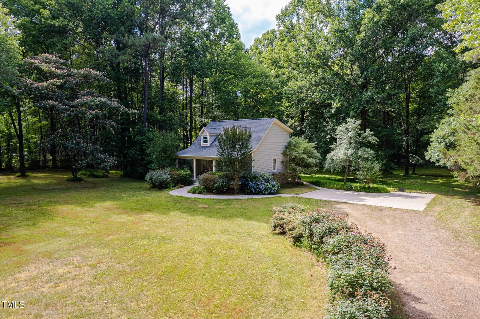 A small beige house with a dark roof is surrounded by trees in a grassy area. The house has a porch and shrubs nearby. A narrow driveway leads to an adjacent paved area. The setting is peaceful and green.