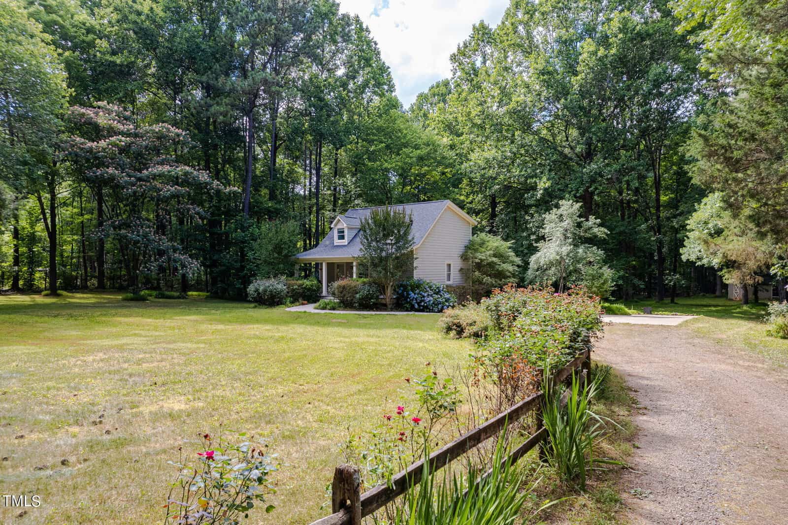 A quaint house with a sloped roof is nestled among tall, lush trees. It has a well-maintained garden with shrubs and flowers. A dirt path lined with a wooden fence leads up to the house, and the foreground features an open grassy area.