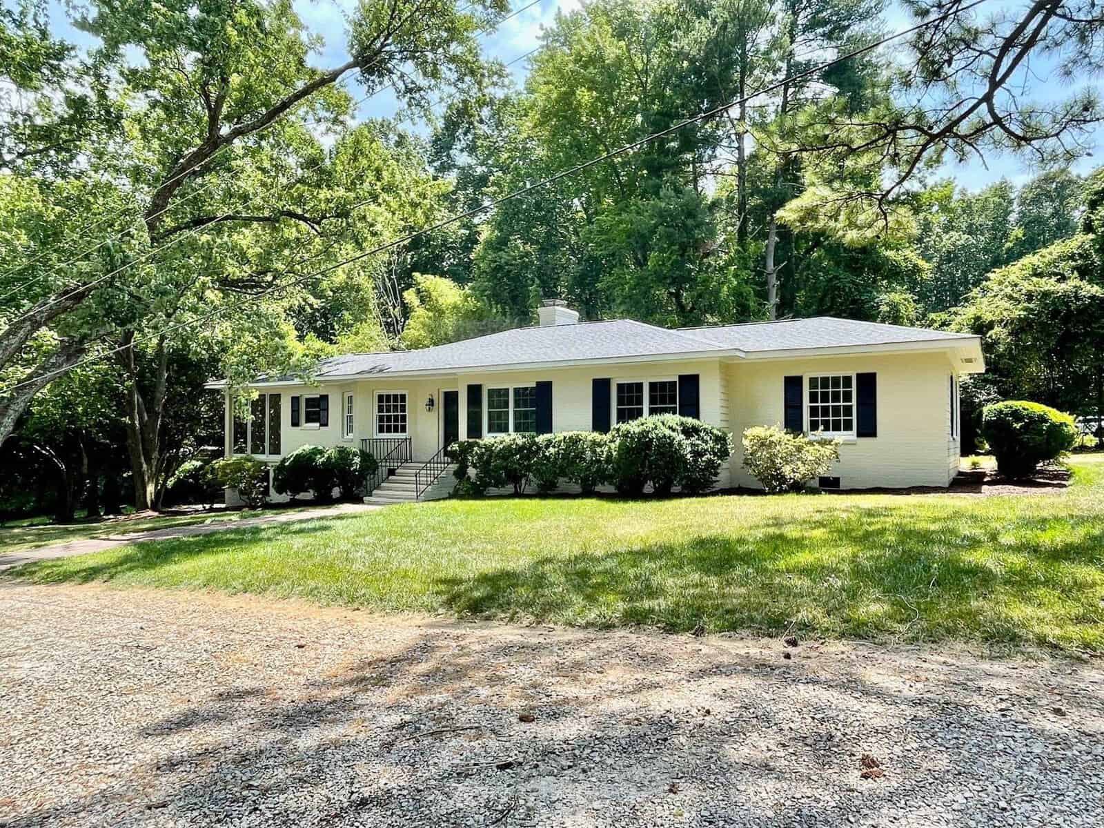 A single-story yellow house with a gray roof sits among trees and shrubs at 645 Lakeview Drive, Henderson NC. It has dark blue shutters, a front porch with steps, and a spacious grassy yard. The gravel driveway in the foreground is surrounded by trees, providing shade and greenery.